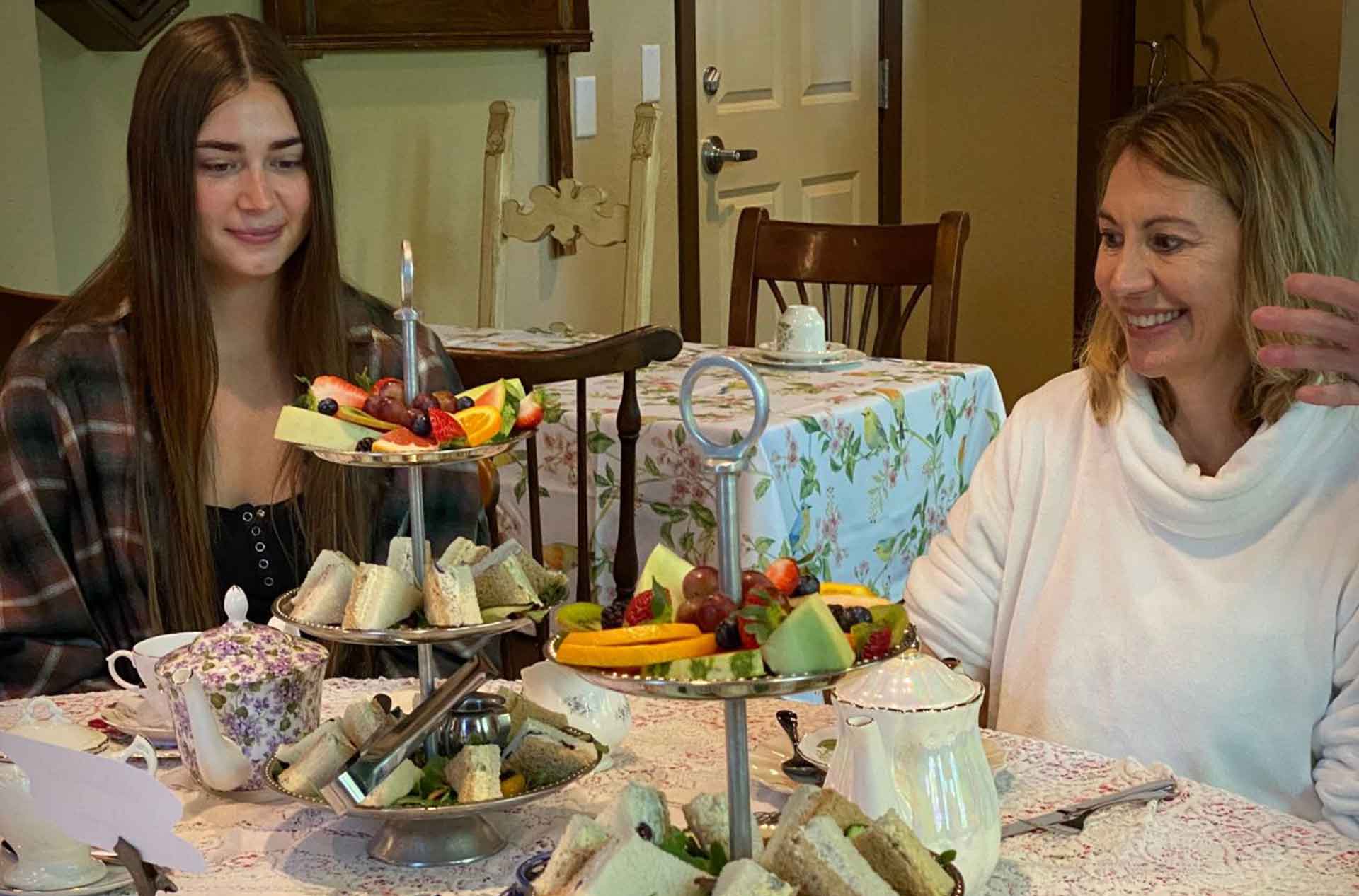 Two people seated at a floral-covered table enjoying afternoon tea with tiered trays of finger sandwiches and fresh fruit in a cozy, vintage-style setting.