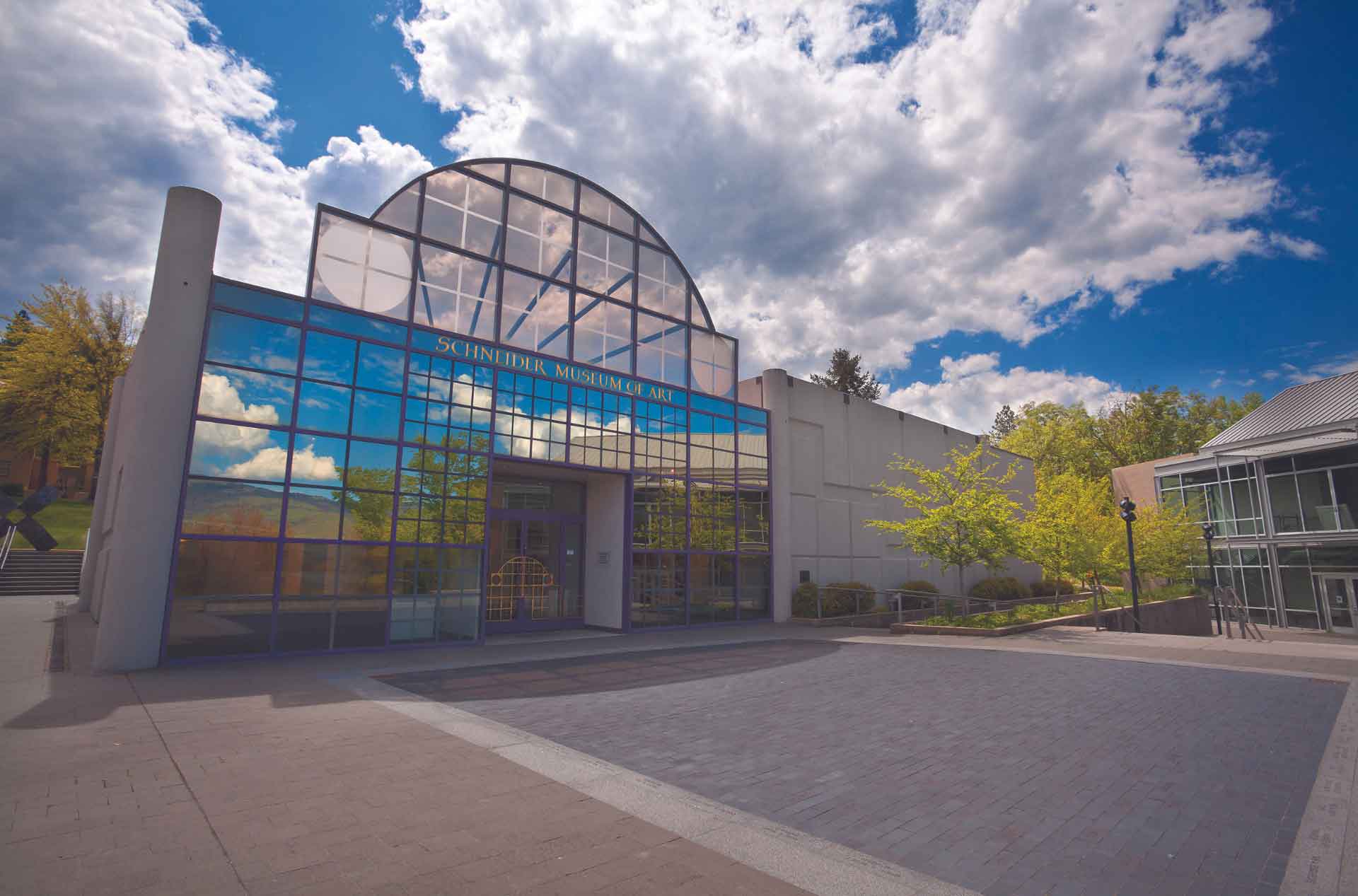 The Schneider Museum of Art's exterior features a modern architectural design with a large glass facade reflecting the blue sky and clouds. The entrance is framed by a semicircular glass arch, and the surrounding area is landscaped with greenery and contemporary structures.
