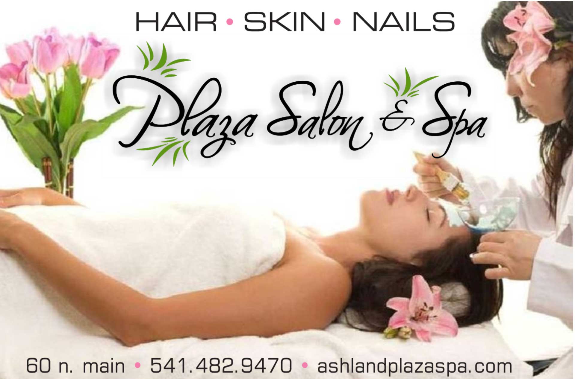 A person is lying on a spa bed covered with a white towel while receiving a facial treatment from a professional applying a mask with a brush. Pink lilies are placed near the person's head, and a vase of pink flowers is in the background. 