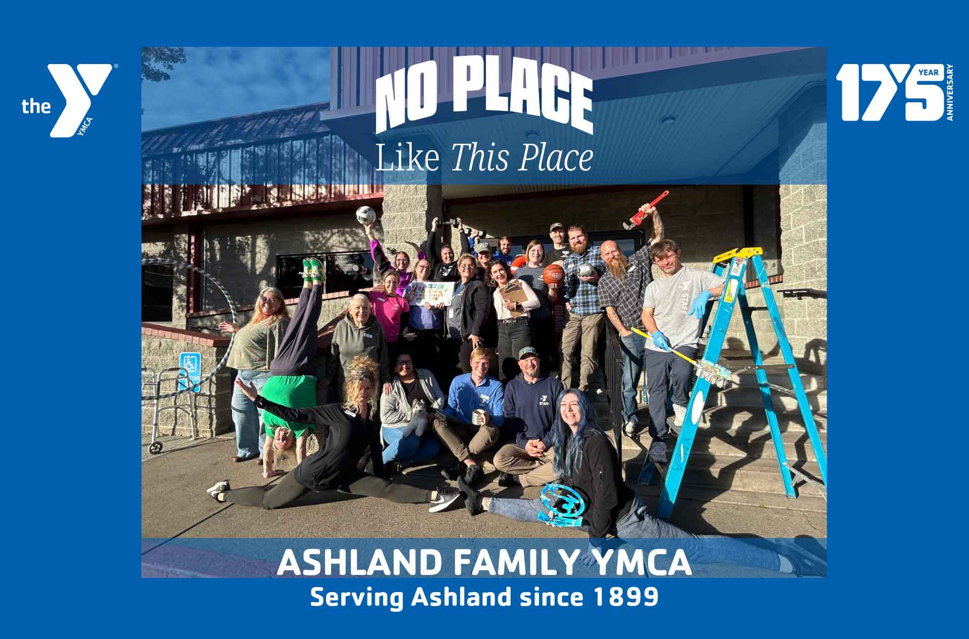 A group of smiling YMCA staff and volunteers posing outside the Ashland Family YMCA building, celebrating together under a banner reading “No Place Like This Place.”