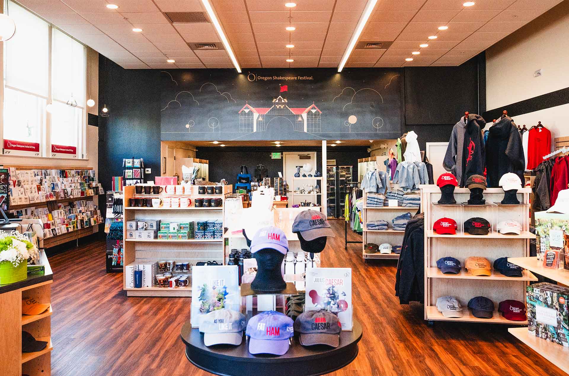 Interior of the Oregon Shakespeare Festival gift shop featuring racks of books, shelves of branded mugs and hats, and displays of hoodies and jackets. A mural of a stylized theater building and trees decorates the back wall above the entrance.