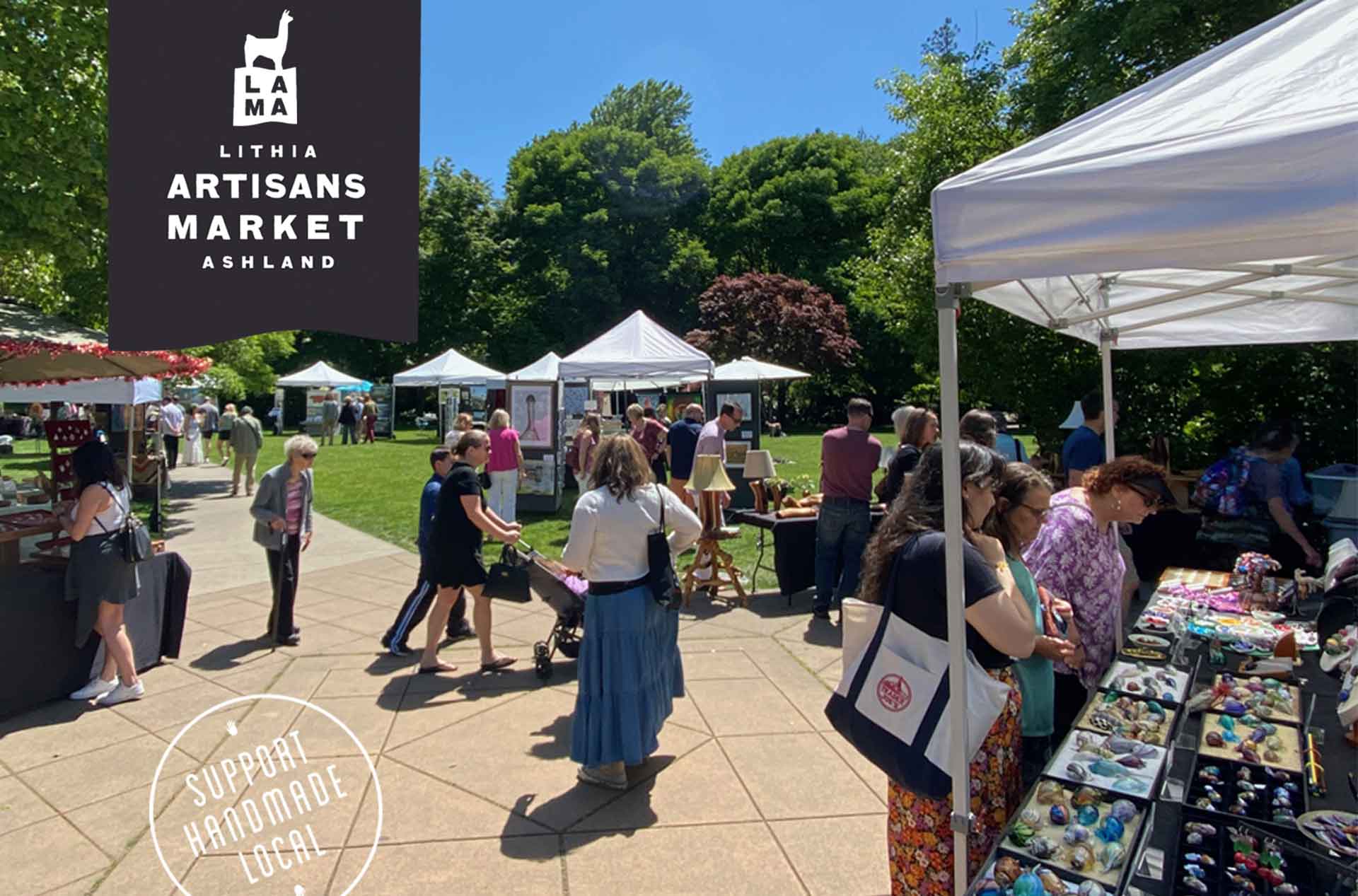 Shoppers browse handmade goods at an outdoor artisan market with white canopy tents set up on a sunny day in a green park.