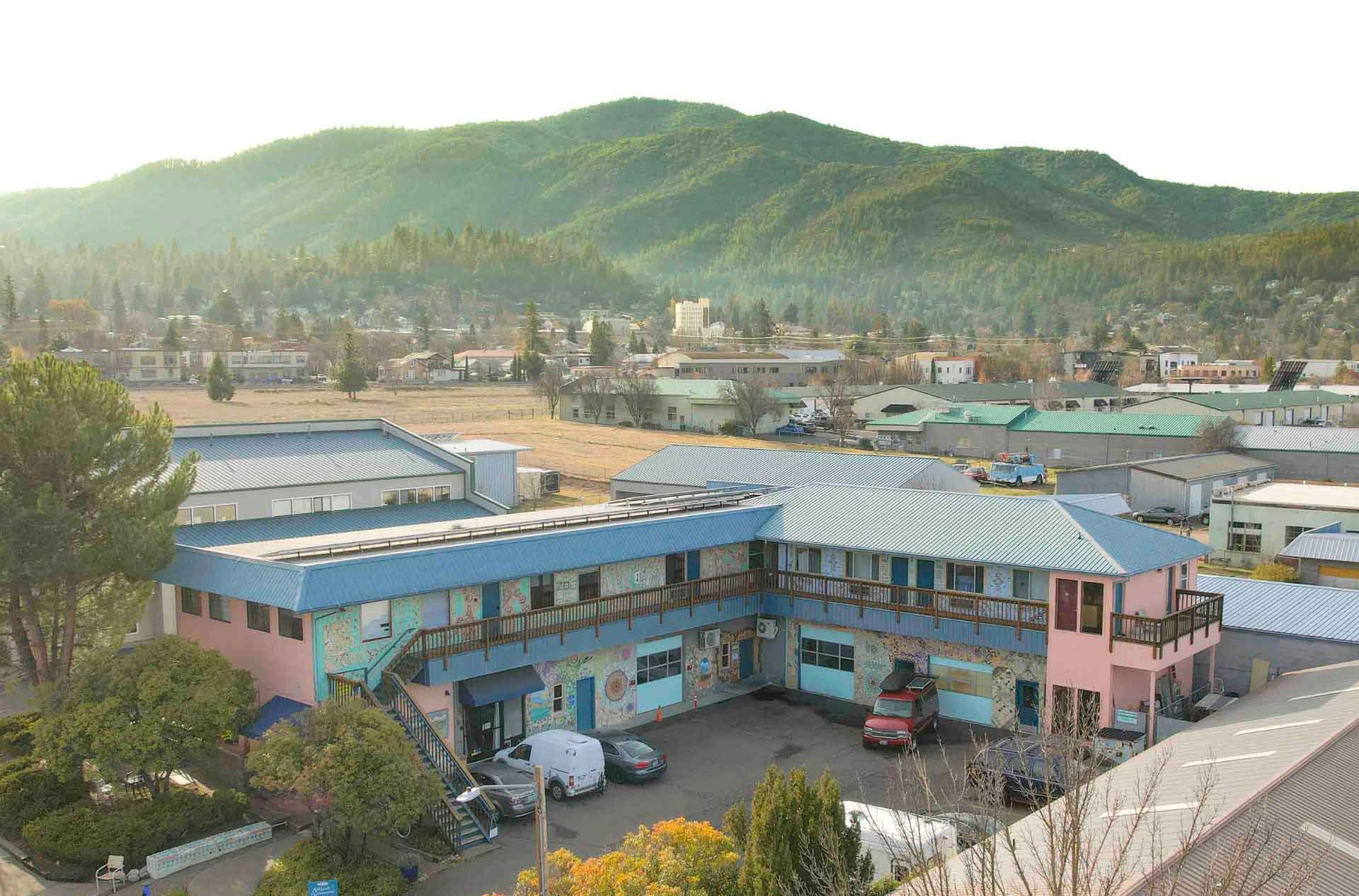 Pastel blue and pink two story building with mosaic art covered walls in a small town, surrounded by forested mountains in soft sunlight.