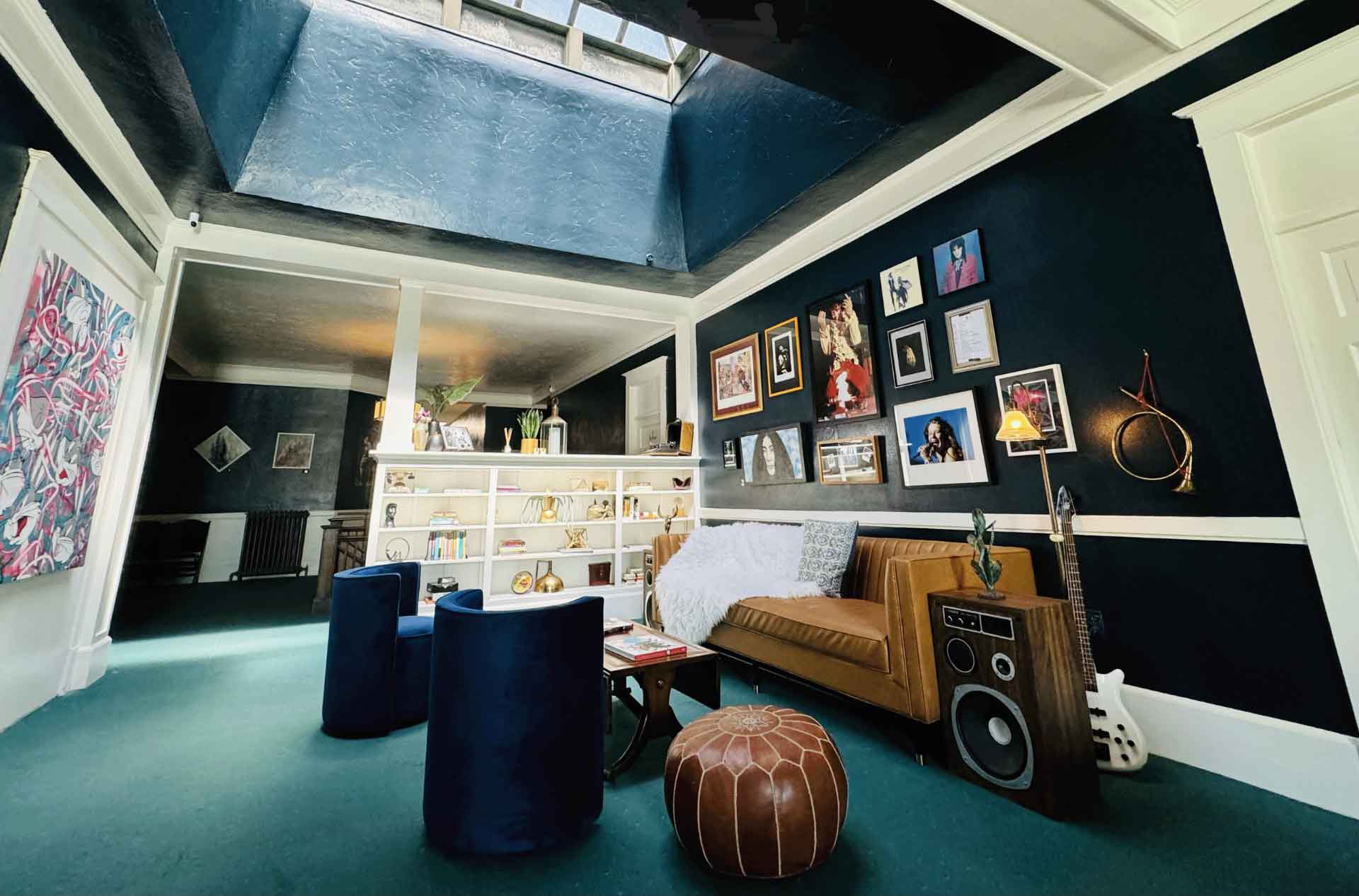Cozy living room with dark teal walls and a large skylight bringing in natural light. A brown leather sofa sits beneath a gallery wall of framed photos, with blue velvet chairs, a large speaker, guitar, and white shelving filled with books and decor.