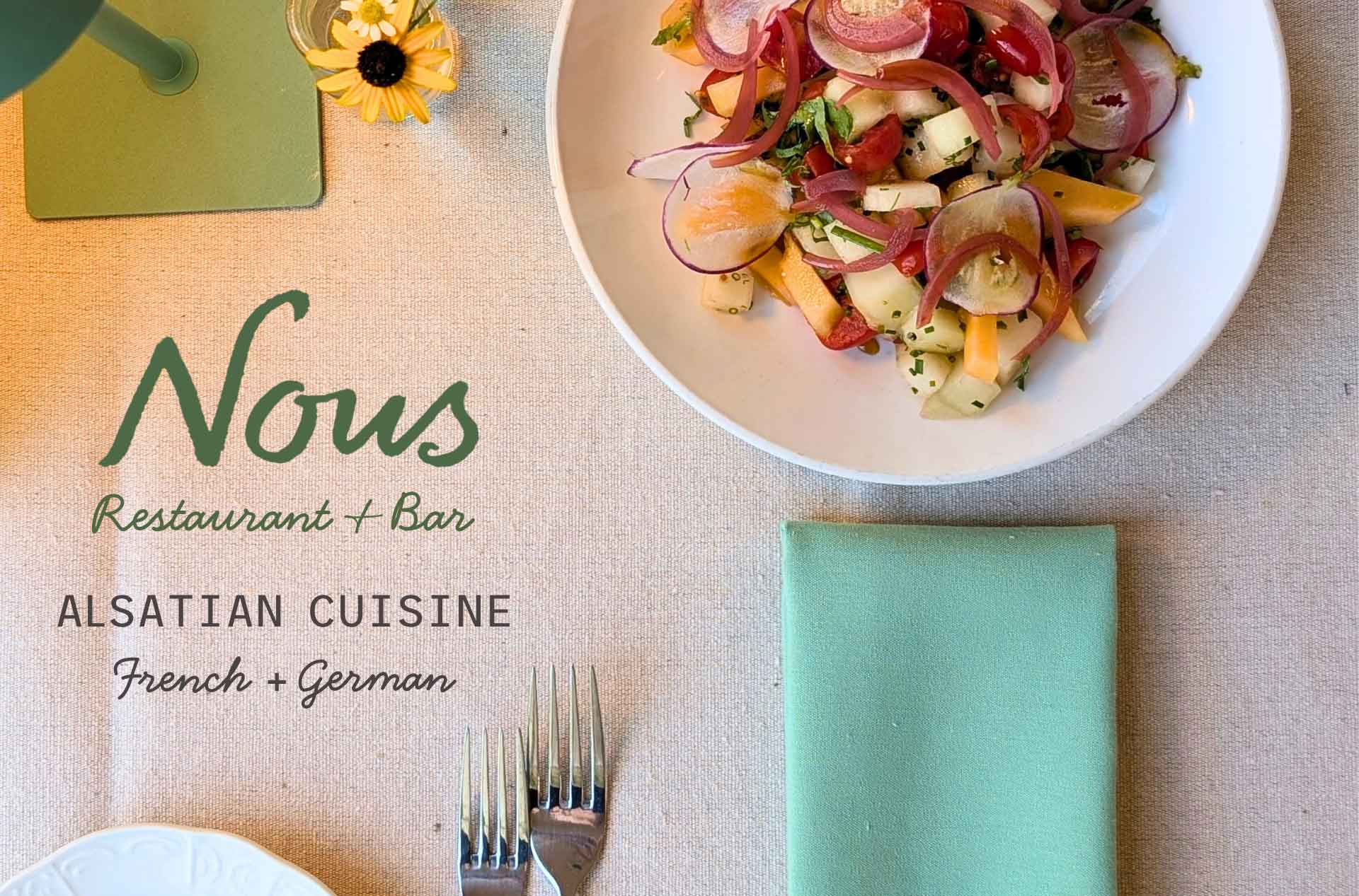 Overhead view of a colorful plated salad with sliced vegetables and herbs on a neatly set table. Text on the left reads “Nous Restaurant + Bar Alsatian Cuisine French + German,” indicating the restaurant and its cuisine style.