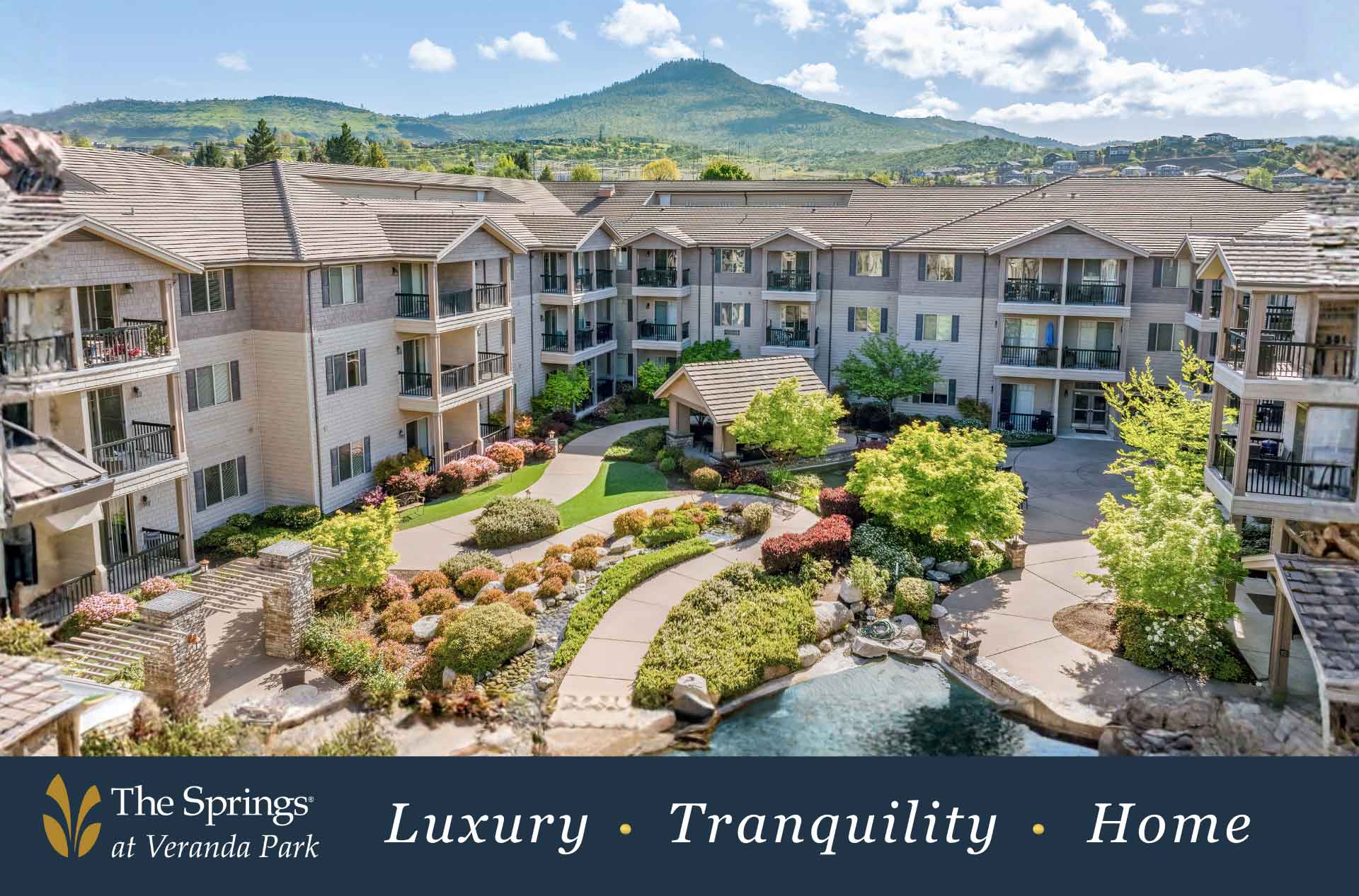 Large apartment complex courtyard with landscaped gardens, winding walkways, and a small pond at the center. Multi story residential buildings with balconies surround the space, with green hills and blue sky in the background and a banner reading "The Springs at Veranda Park Luxury Tranquility Home".