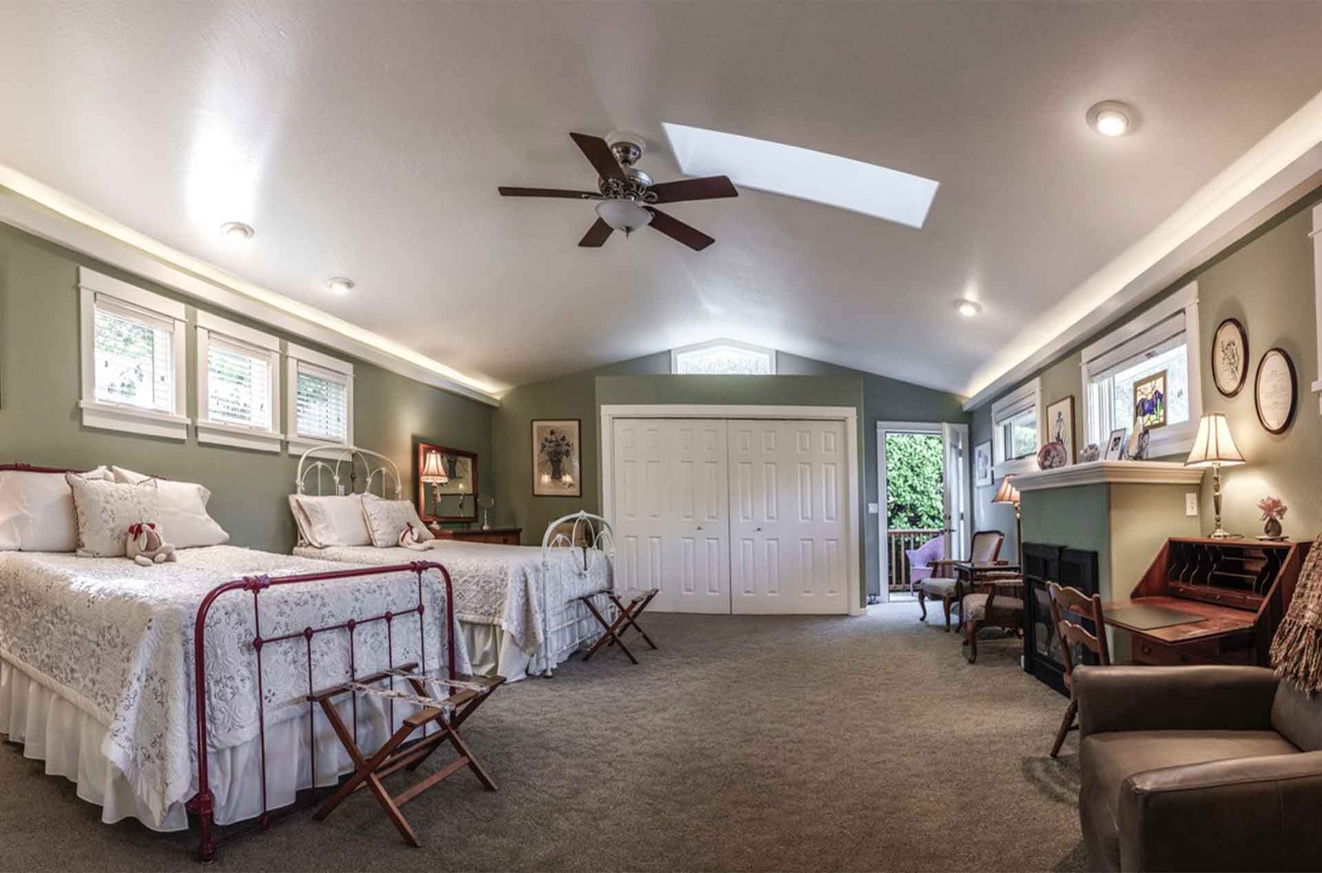Spacious bedroom with two twin beds dressed in white quilts, set against green walls with multiple windows. A vaulted ceiling with a skylight and ceiling fan brightens the room, while a fireplace, desk, and seating area add a cozy feel.