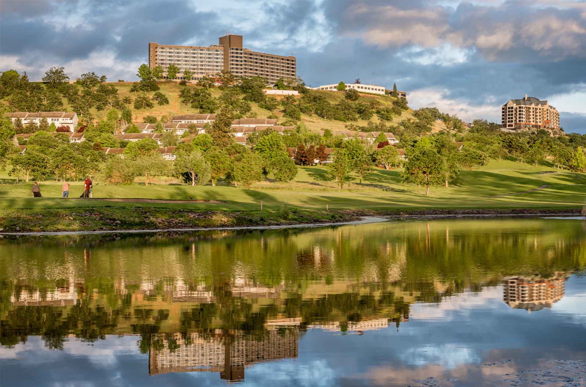 Large buildings sit on a sunlit hillside overlooking a green golf course and calm pond. The water reflects the buildings, trees, and dramatic clouds in the evening sky.