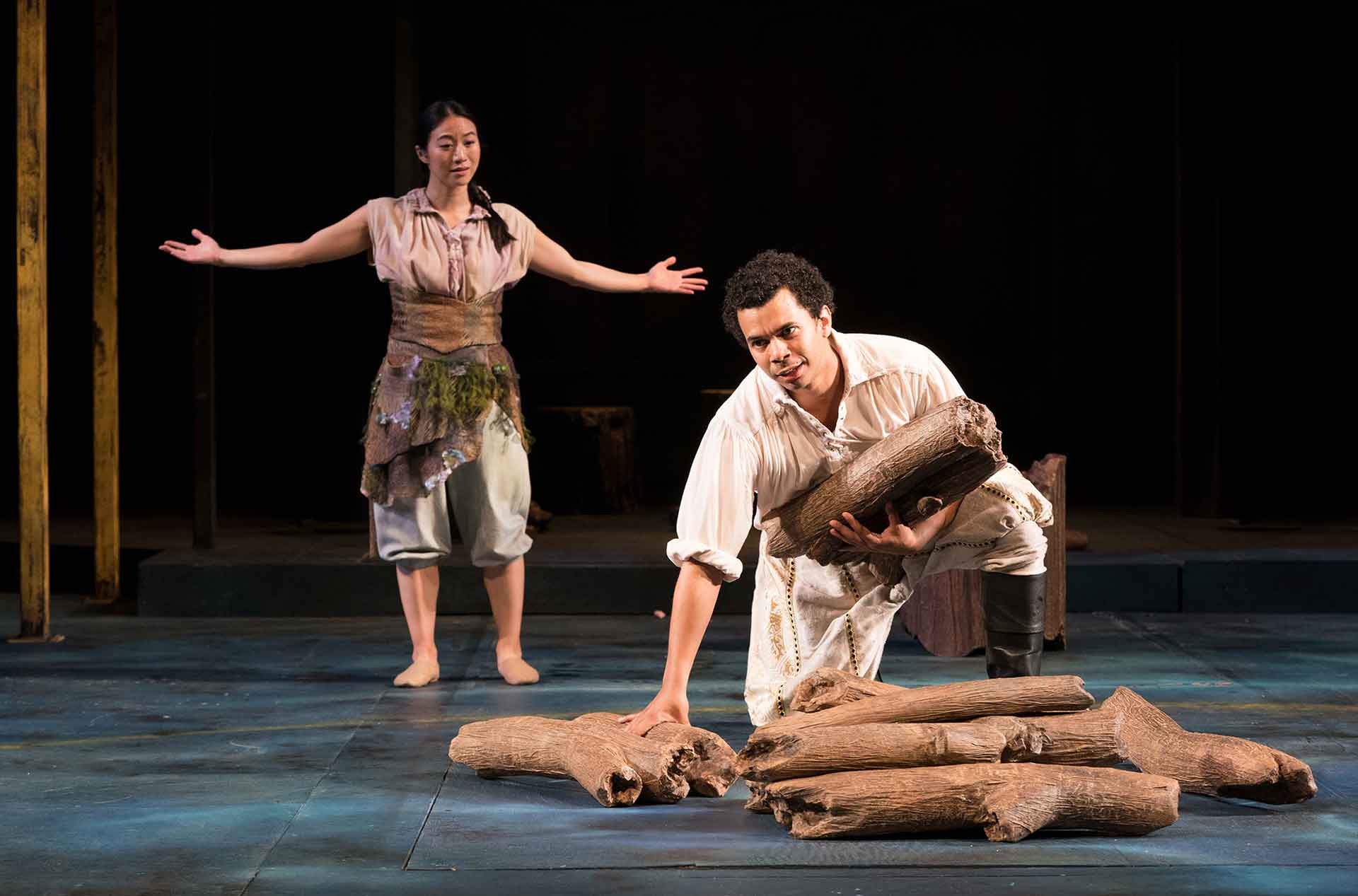 Woman with arms spread standing behind a man stooping to pick up wood.