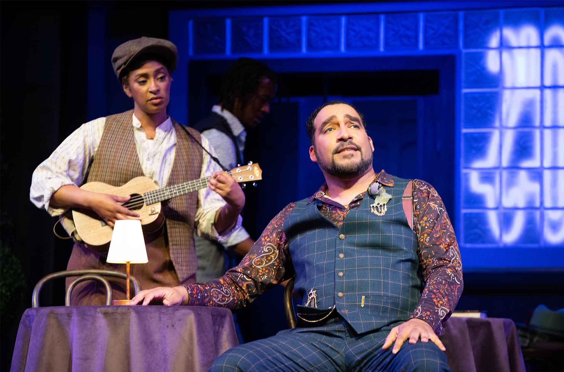 A person playing a guitar to another seated at a table.