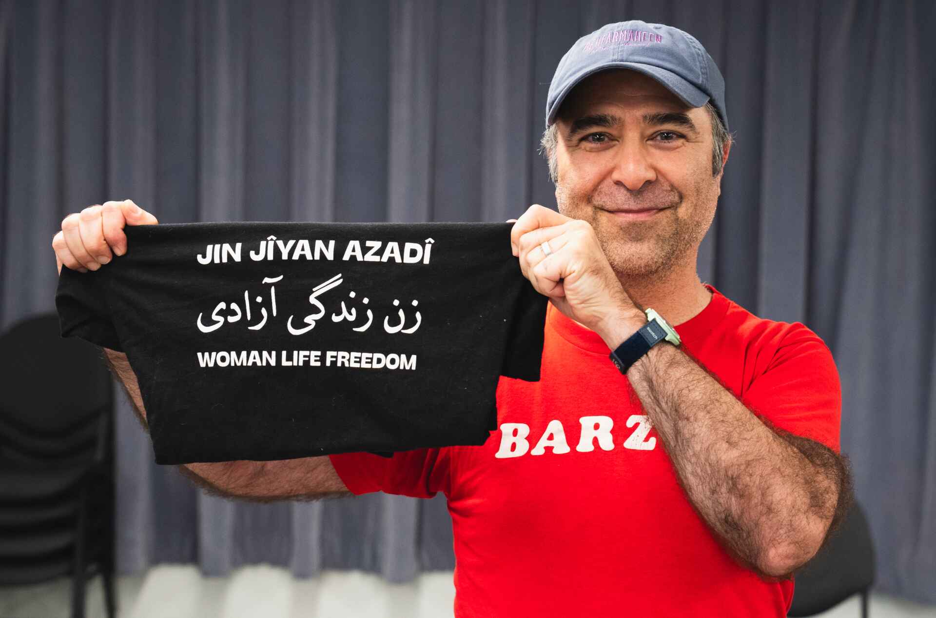 A person in a red shirt and gray cap is holding a black T-shirt with the text "JIN JIYAN AZADÎ" in a foreign language and "WOMAN LIFE FREEDOM" in English.