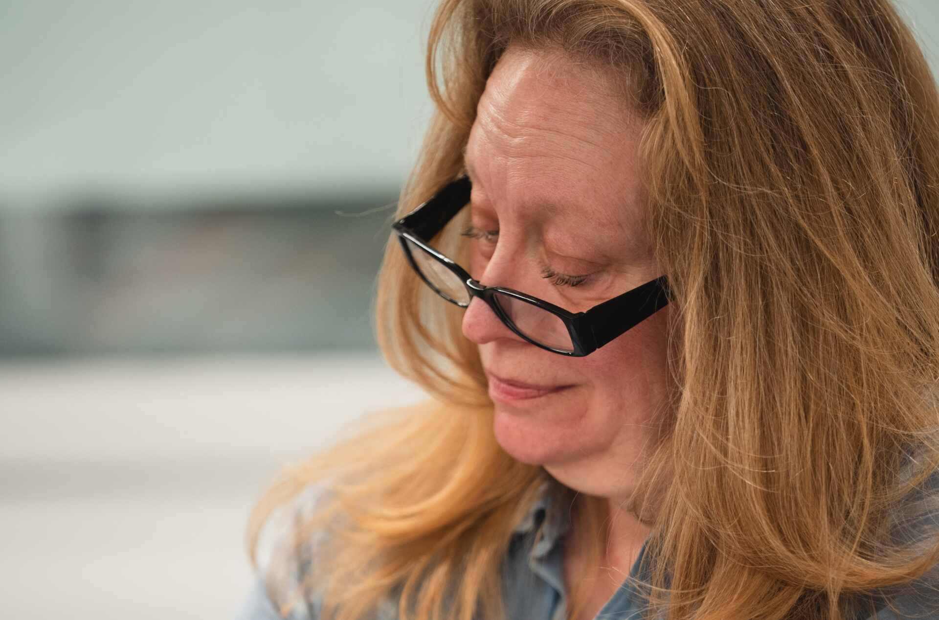 A person with long blonde hair wearing glasses is sitting and writing something on a piece of paper. They are dressed in a blue plaid shirt and is focused on her task. They have a green wristwatch on their left wrist.