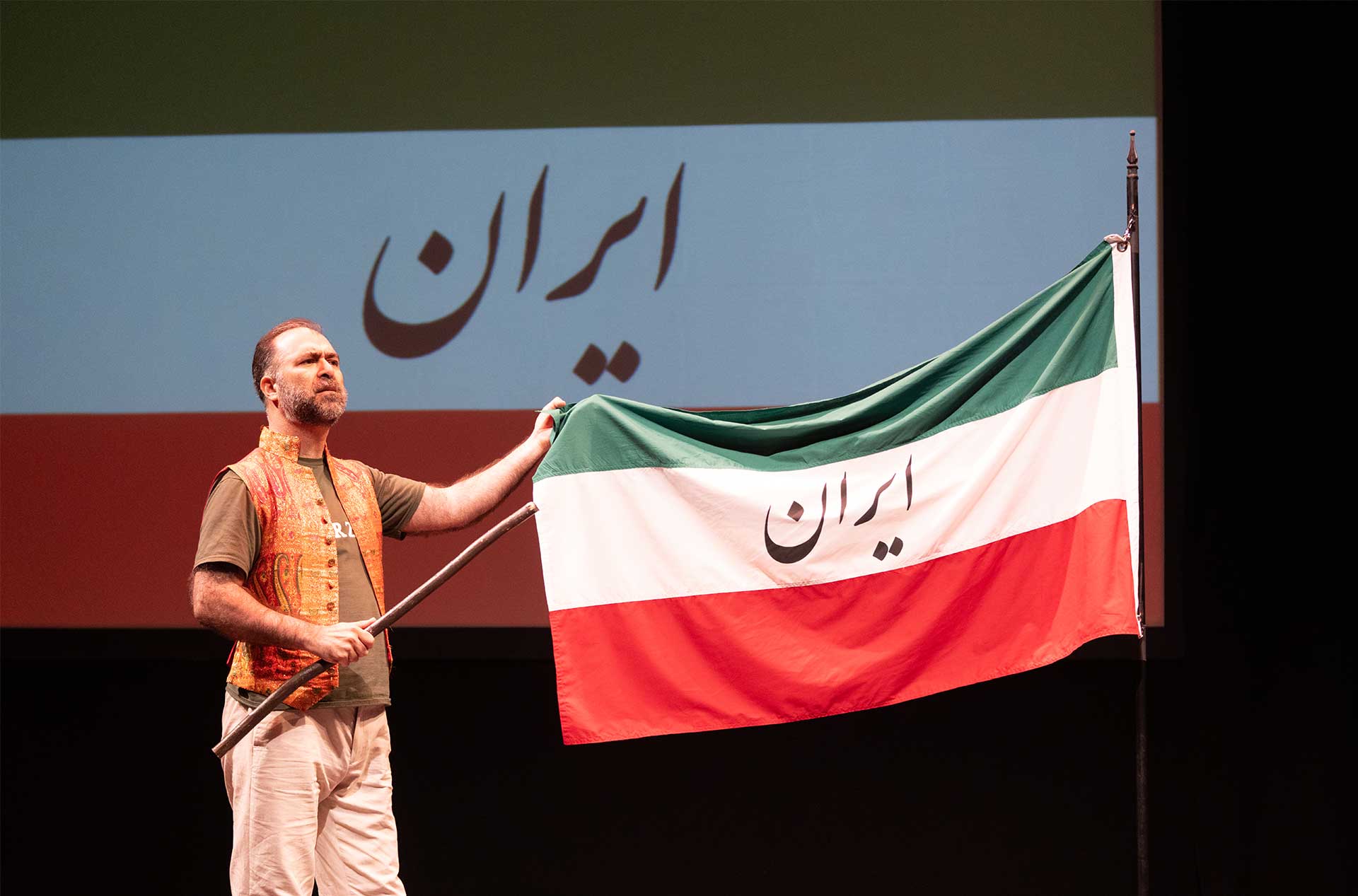 A person holds an Iranian flag on stage, with the same flag projected on the screen behind them. The flag displays the word "Iran" in Persian script.