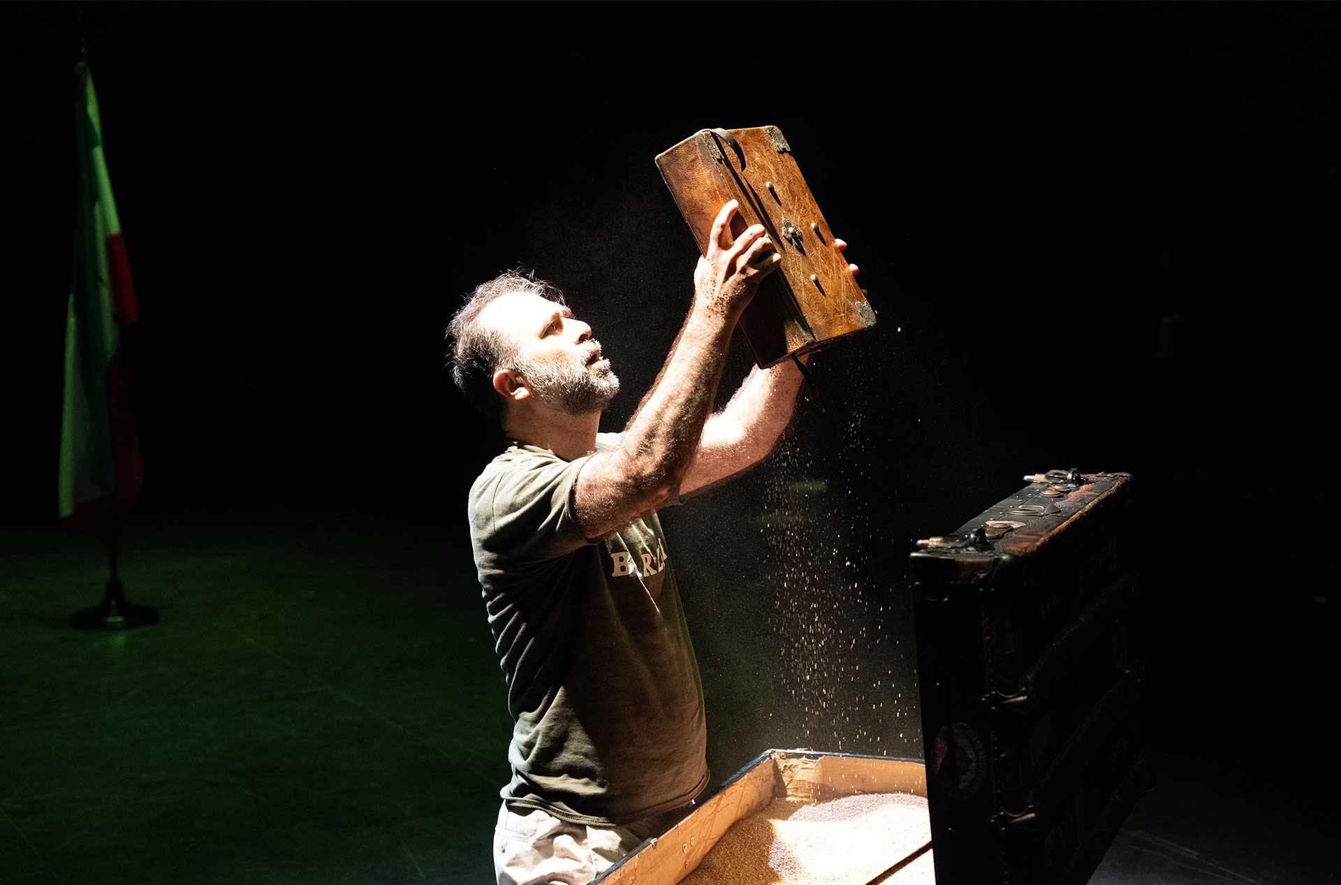A person in a spotlight holds an old book above a wooden box, with particles scattering in the light, while a flag is visible in the background.