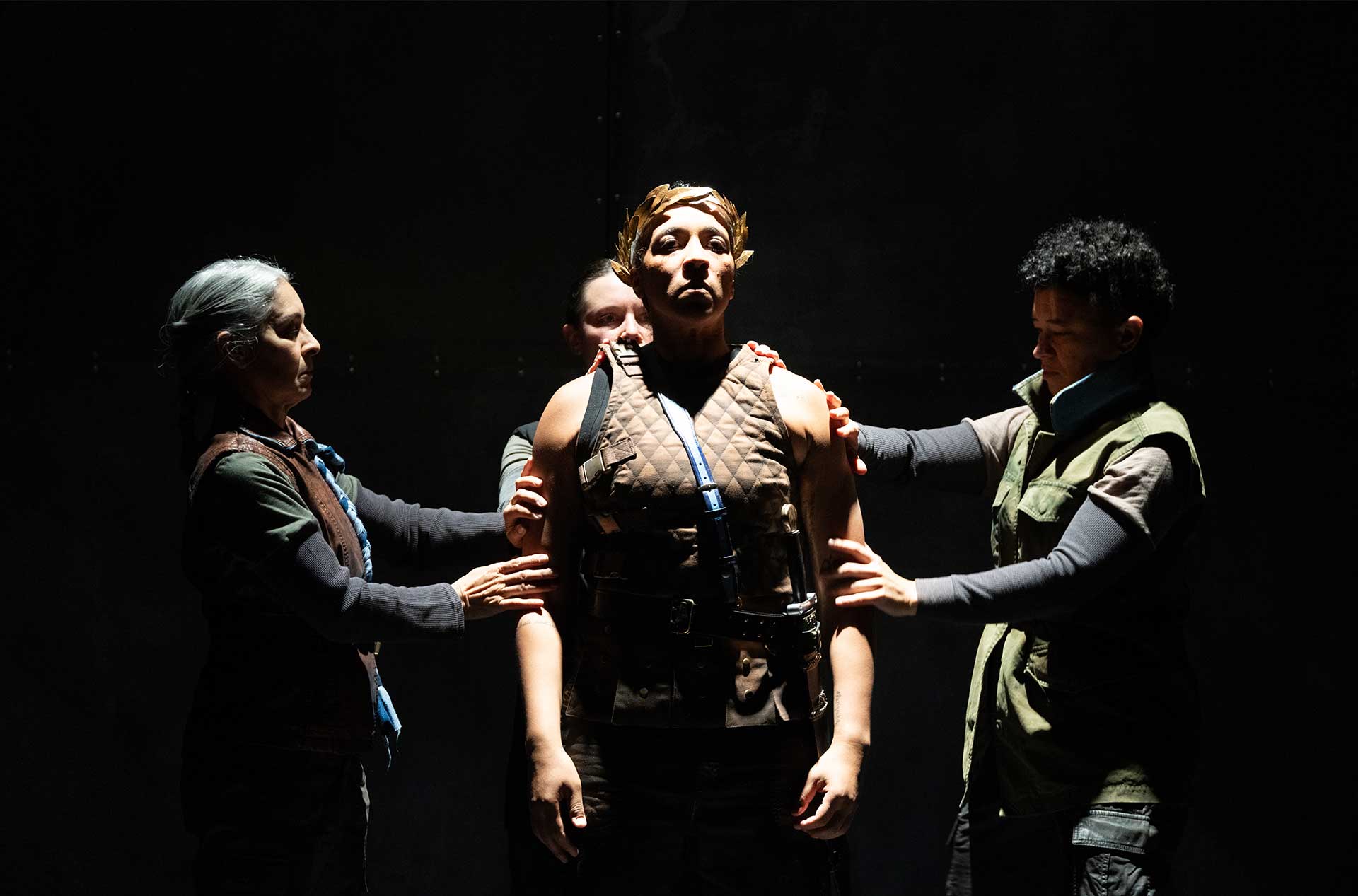 A central figure wearing a laurel crown and a brown quilted vest stands with a solemn expression while three others stand touching them. The focused lighting emphasizes the importance of this character in the scene.