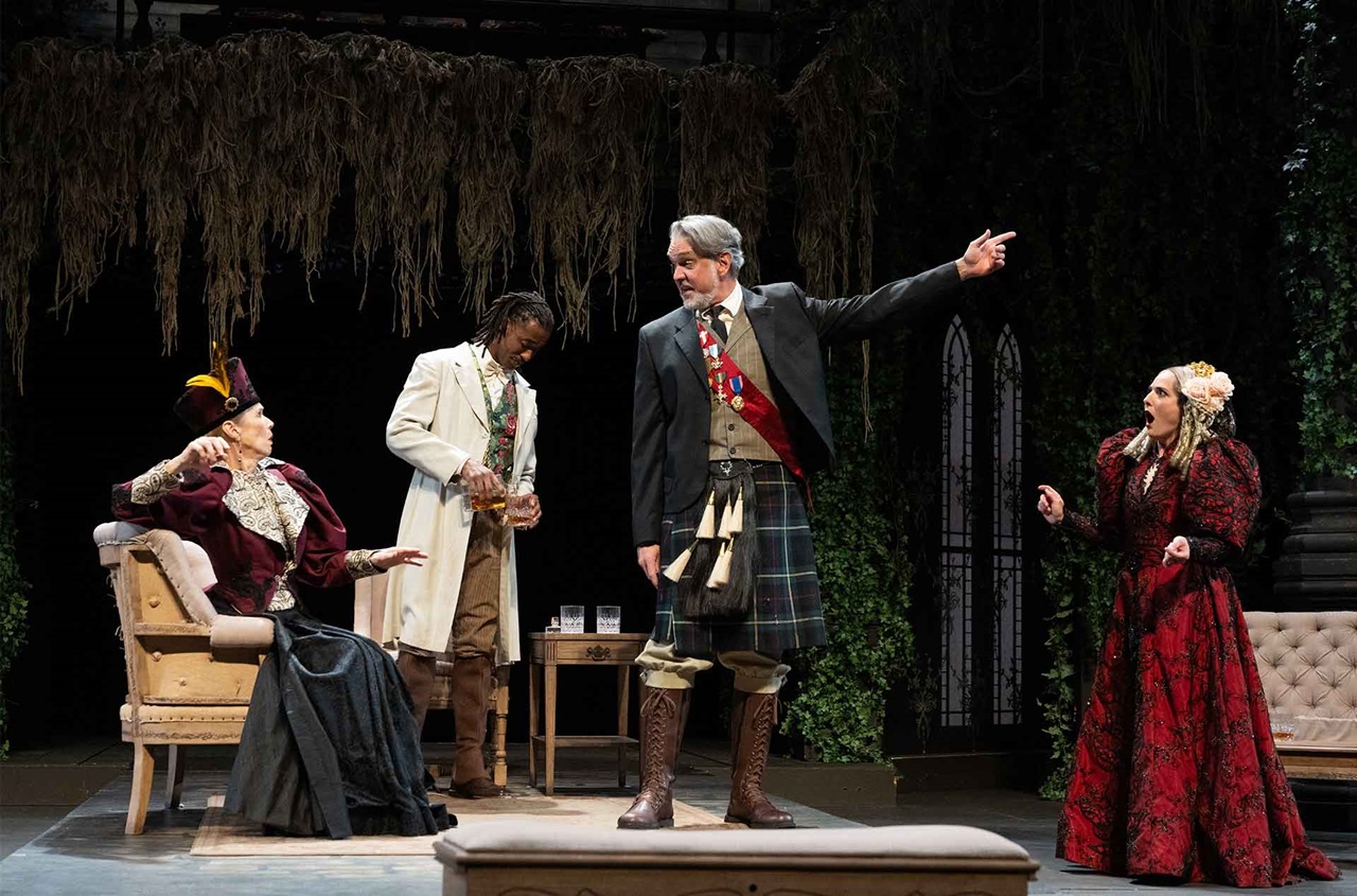 A person wearing a kilt gesturing two the right, a person sitting in a chair and another standing watching, a fourth person looking at the ground.