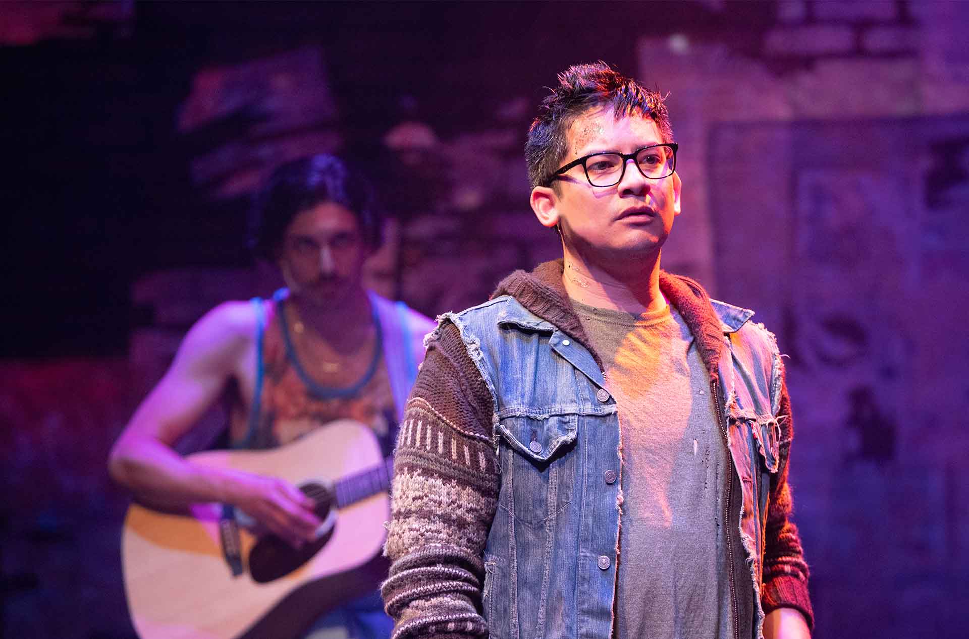 A person in a denim jacket stands with a solemn look in the foreground, while another person in the background, slightly out of focus, plays a guitar.