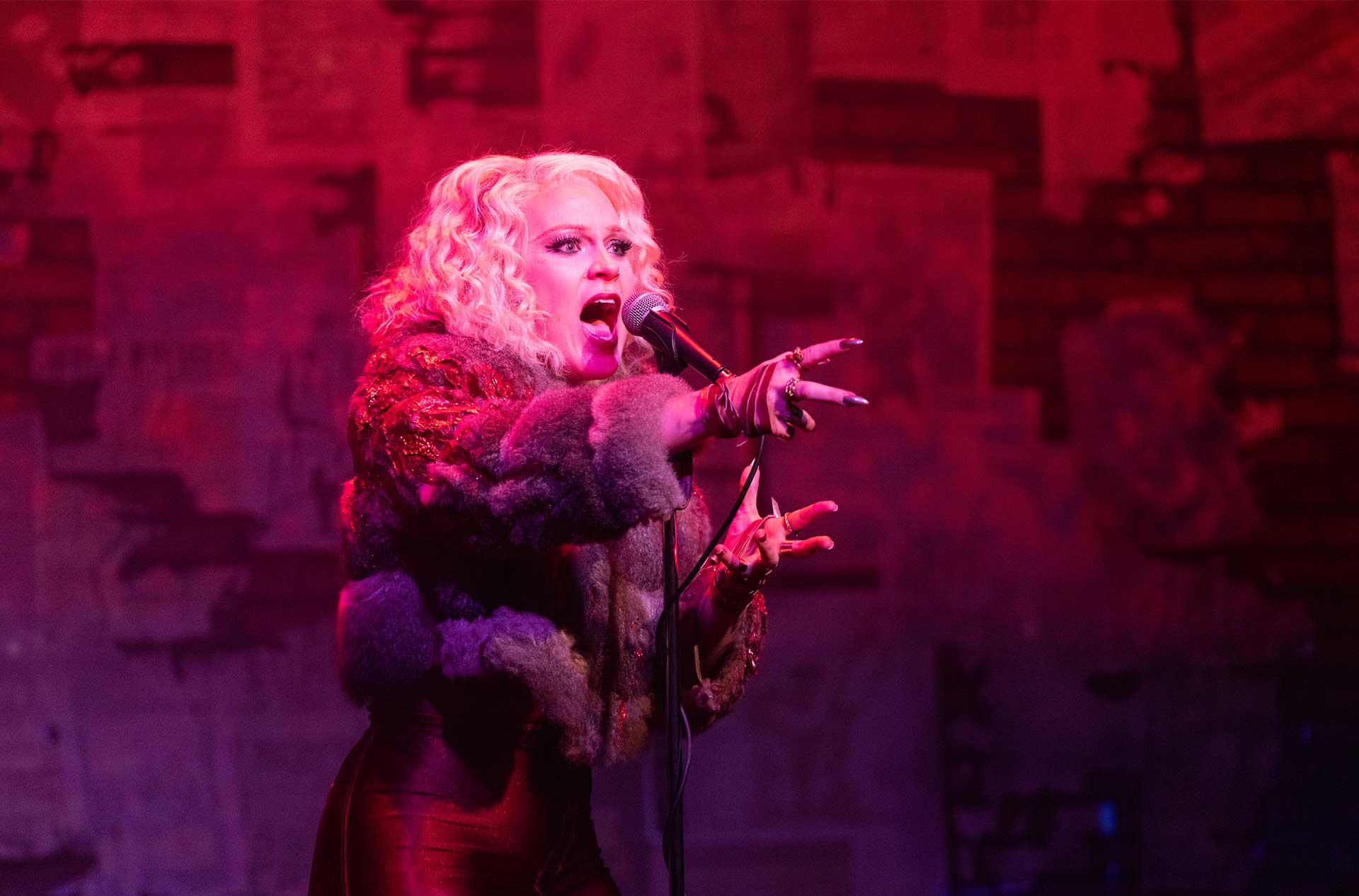 A person with blonde curly hair, wearing a fur coat and red outfit, is passionately singing into a microphone against a red-lit background.