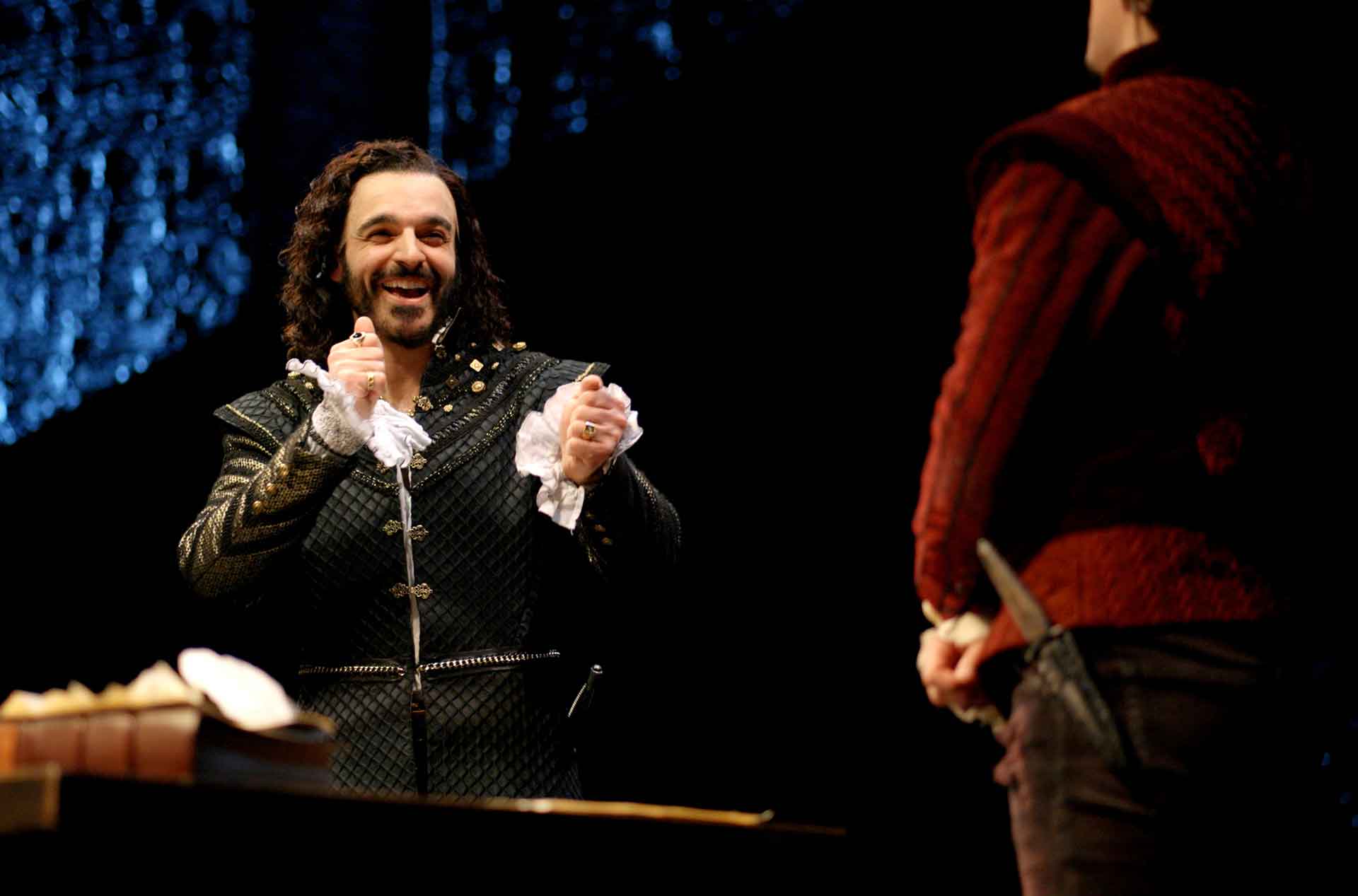 A person dressed in a dark, ornate costume with ruffled sleeves is smiling and clapping, facing another individual in a red outfit who is holding a dagger behind their back. 