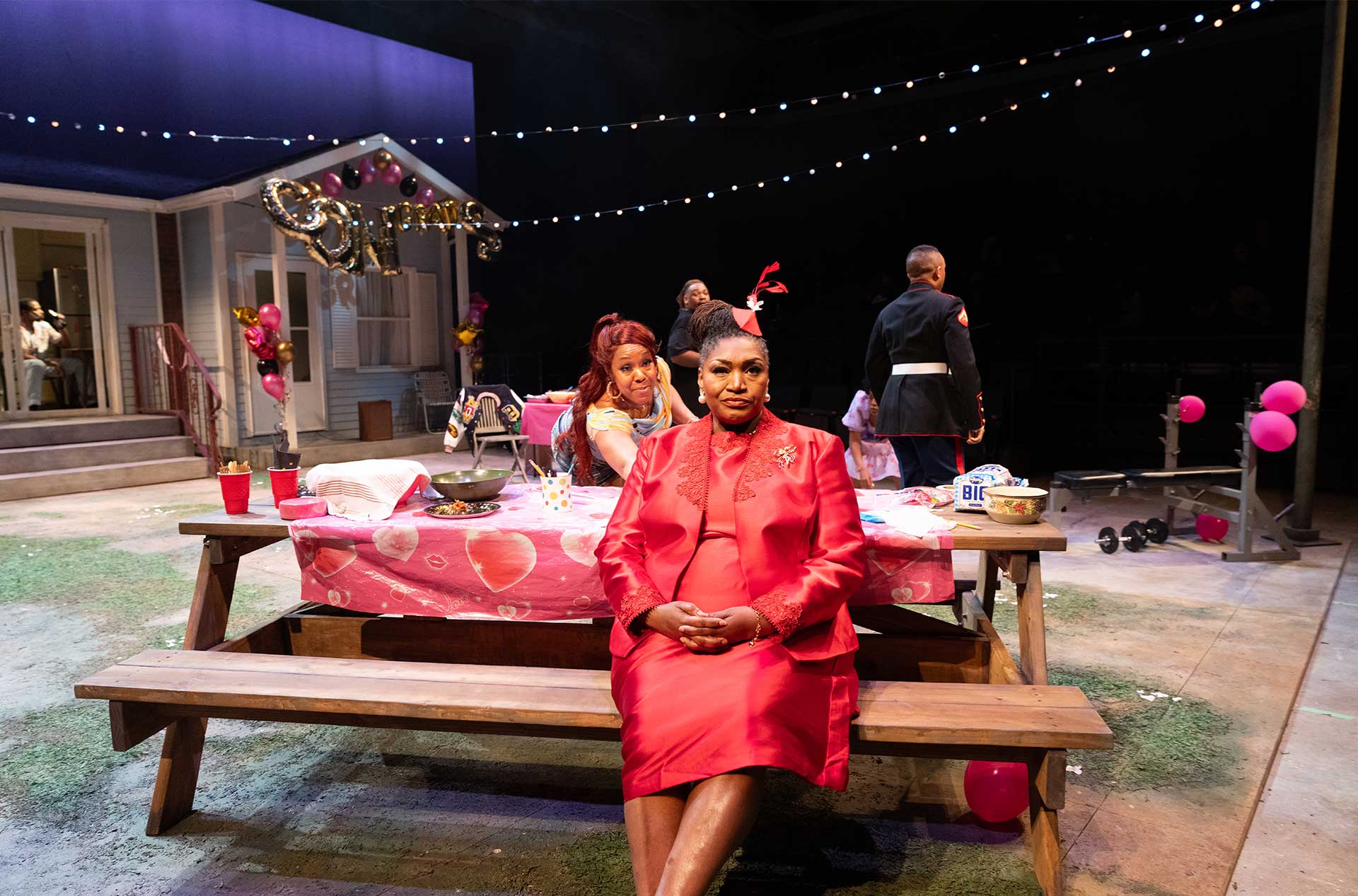 A person in a bright red suit and hat sits calmly at a picnic table covered in a pink heart-themed tablecloth, while others bustle around during an outdoor party scene.