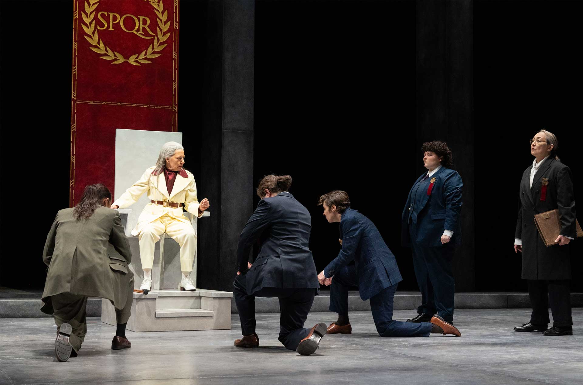 A character in a white suit sits on a modern throne under a red banner that reads "SPQR," as several figures kneel before them while others stand nearby in formal attire.