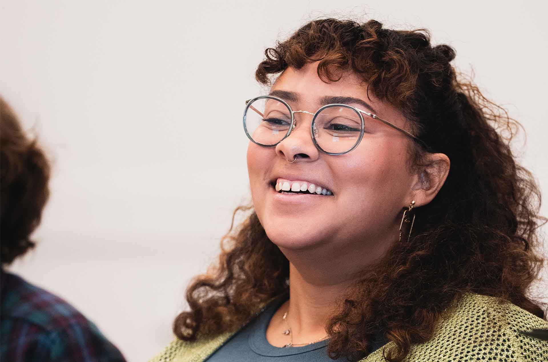 A person with curly hair, glasses, and a nose ring smiles warmly. They wear a green knit cardigan over a blue shirt.