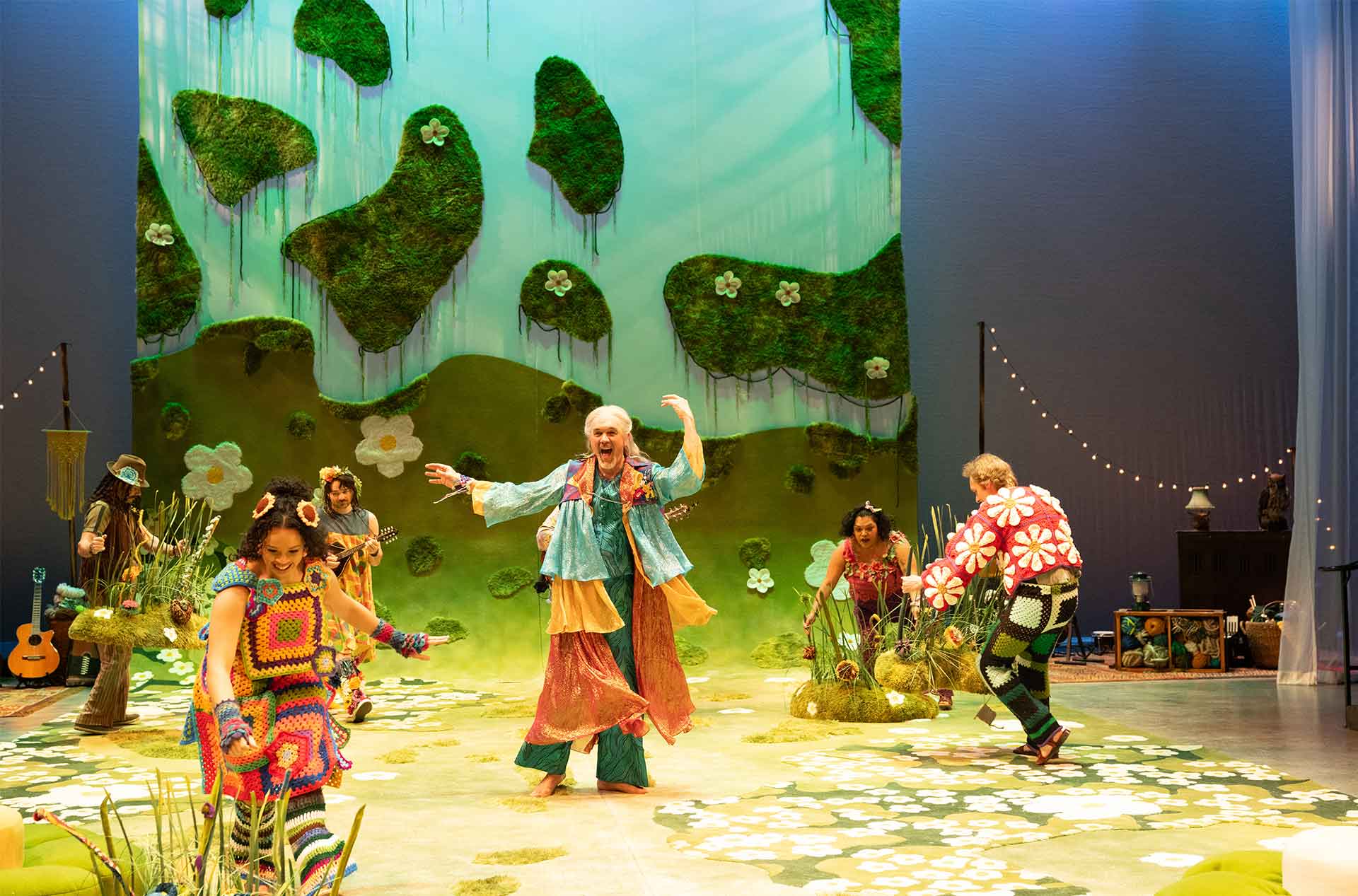 Performers in vibrant crochet costumes dance joyfully on a flower-filled stage, with one leading in front of a backdrop of green hills and felted flowers.