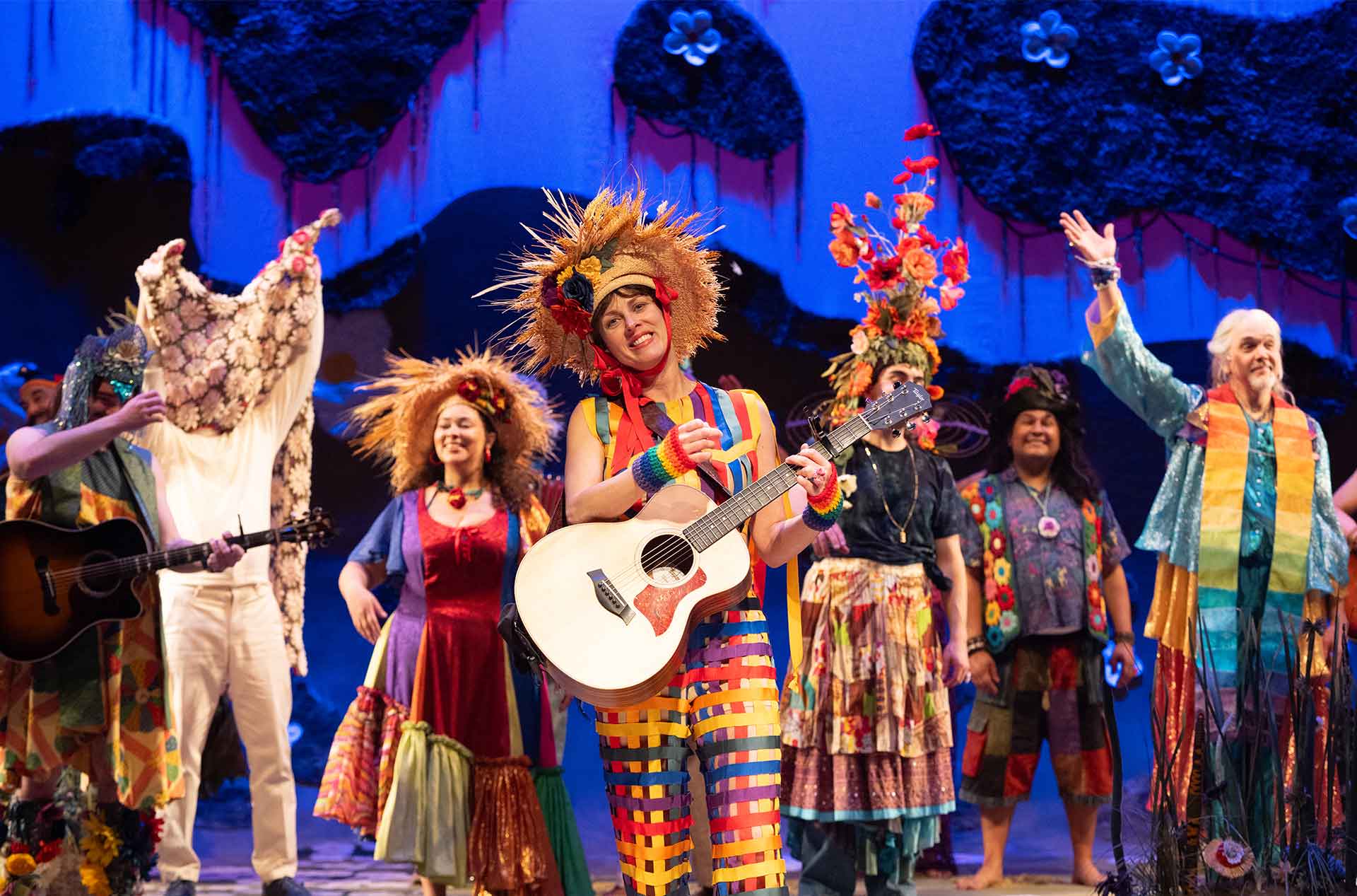 Cast members in vivid, multicolored costumes with flower and ribbon headpieces smile and perform beneath a dreamy blue and pink-lit backdrop with hanging moss and oversized blossoms.