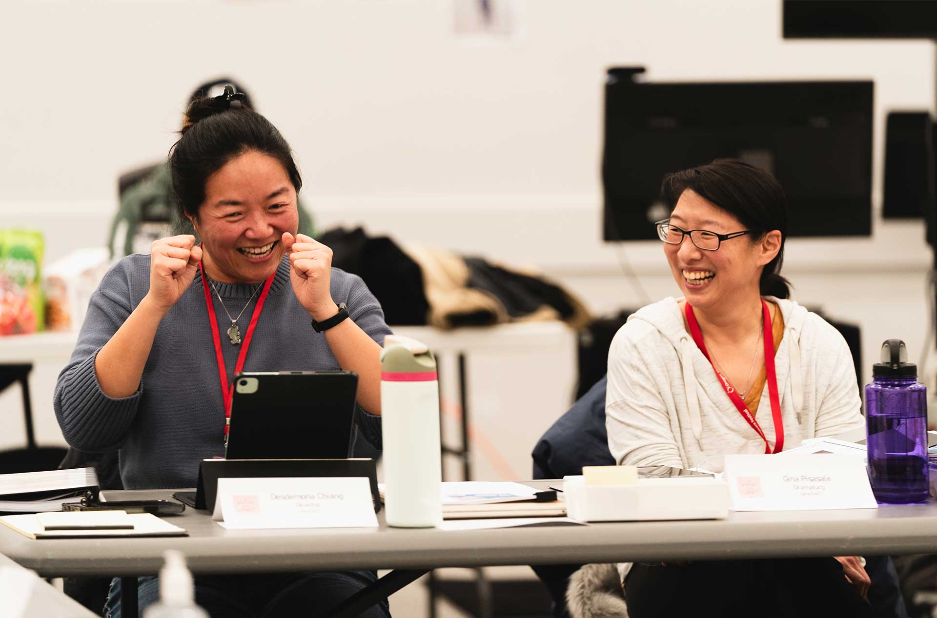 Two individuals seated at a table share a laugh, one gesturing enthusiastically while the other smiles with a relaxed posture. Papers, tablets, and name cards are visible on the table.