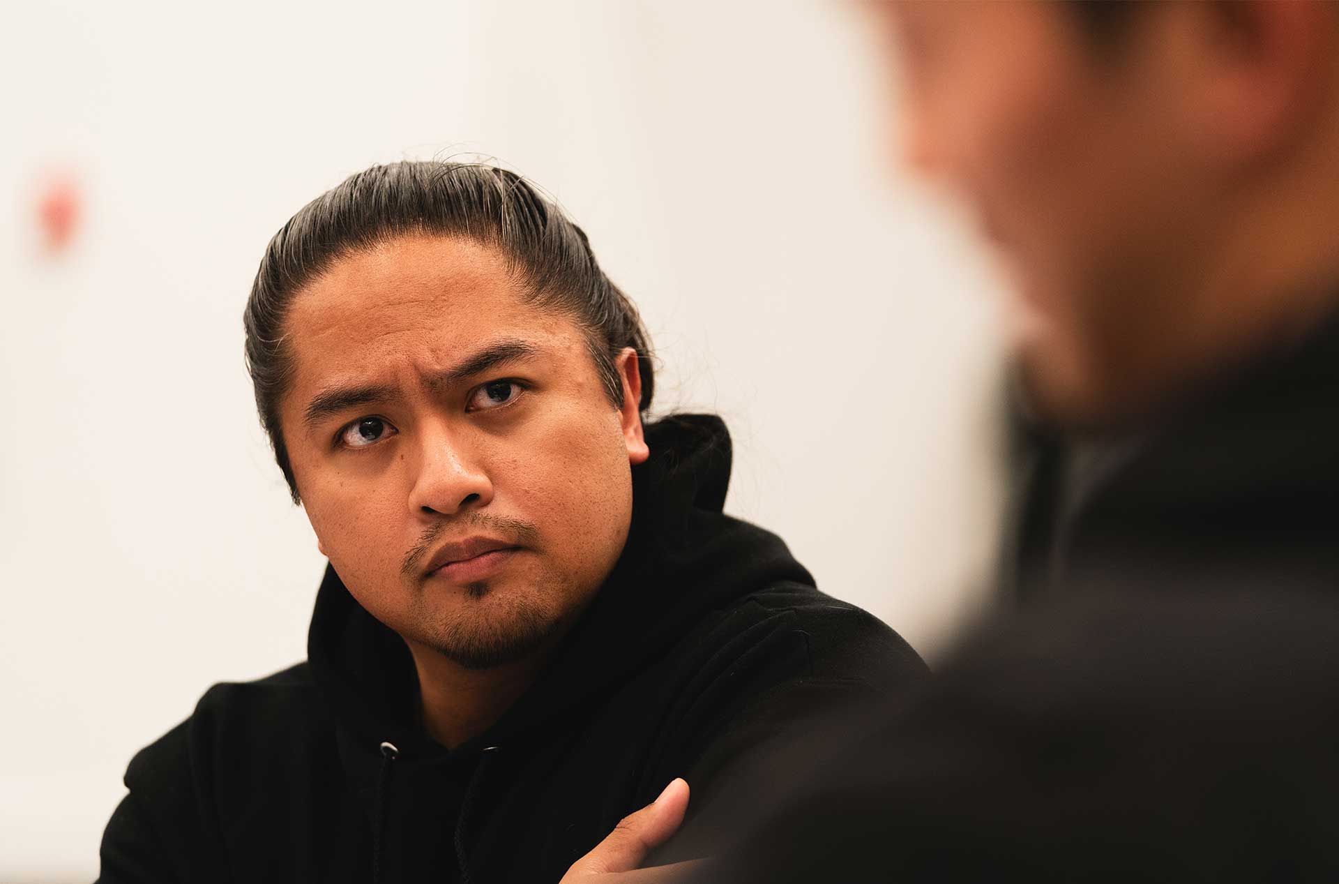 A person with a serious expression sits with their arms crossed, engaging in a conversation with someone whose back is to the camera. The room is softly lit, with blurred details in the background.