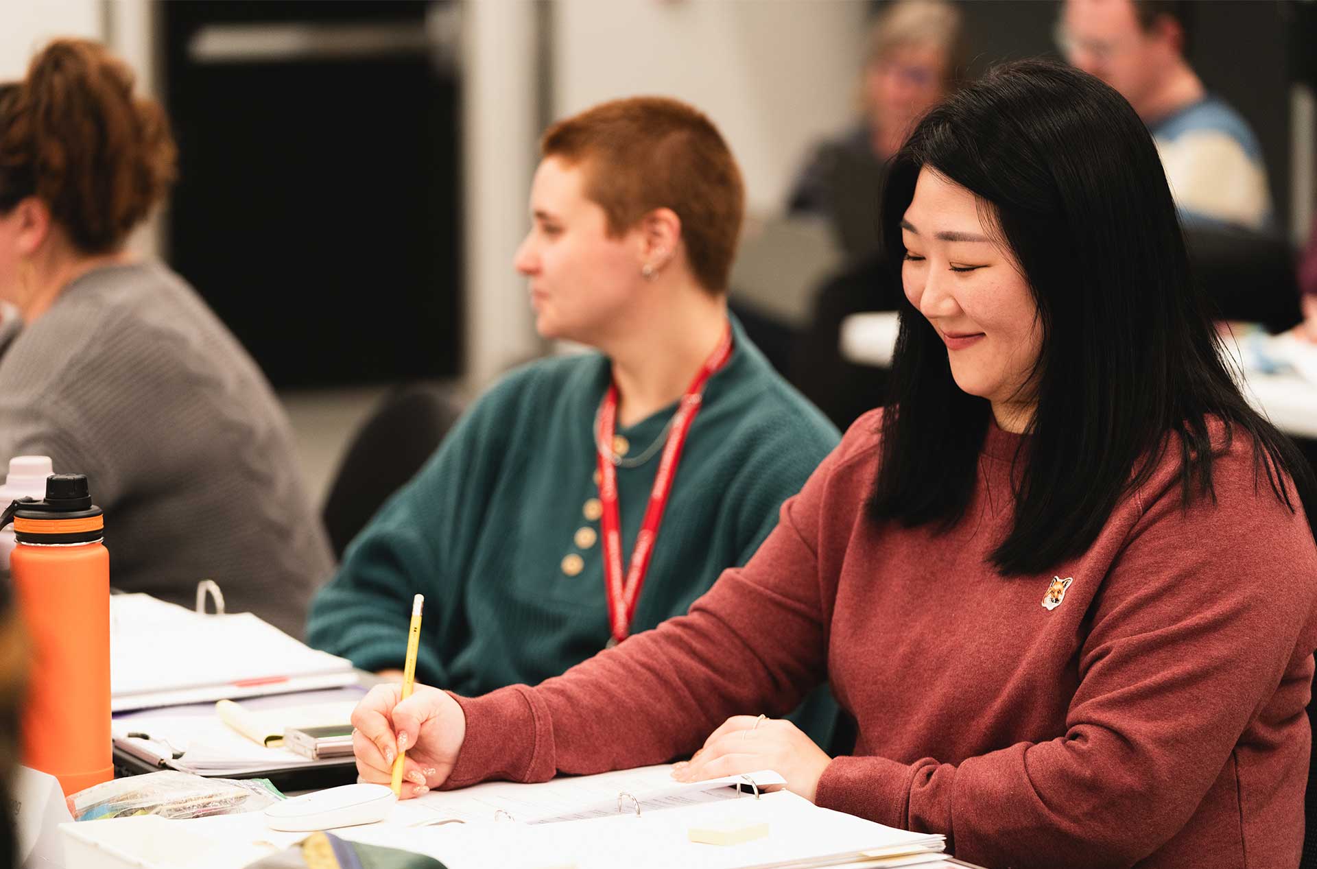 Two people seated at the table focus on their notes, one writing with a pencil while the other listens. The rehearsal atmosphere is collaborative, with materials spread out on the table.