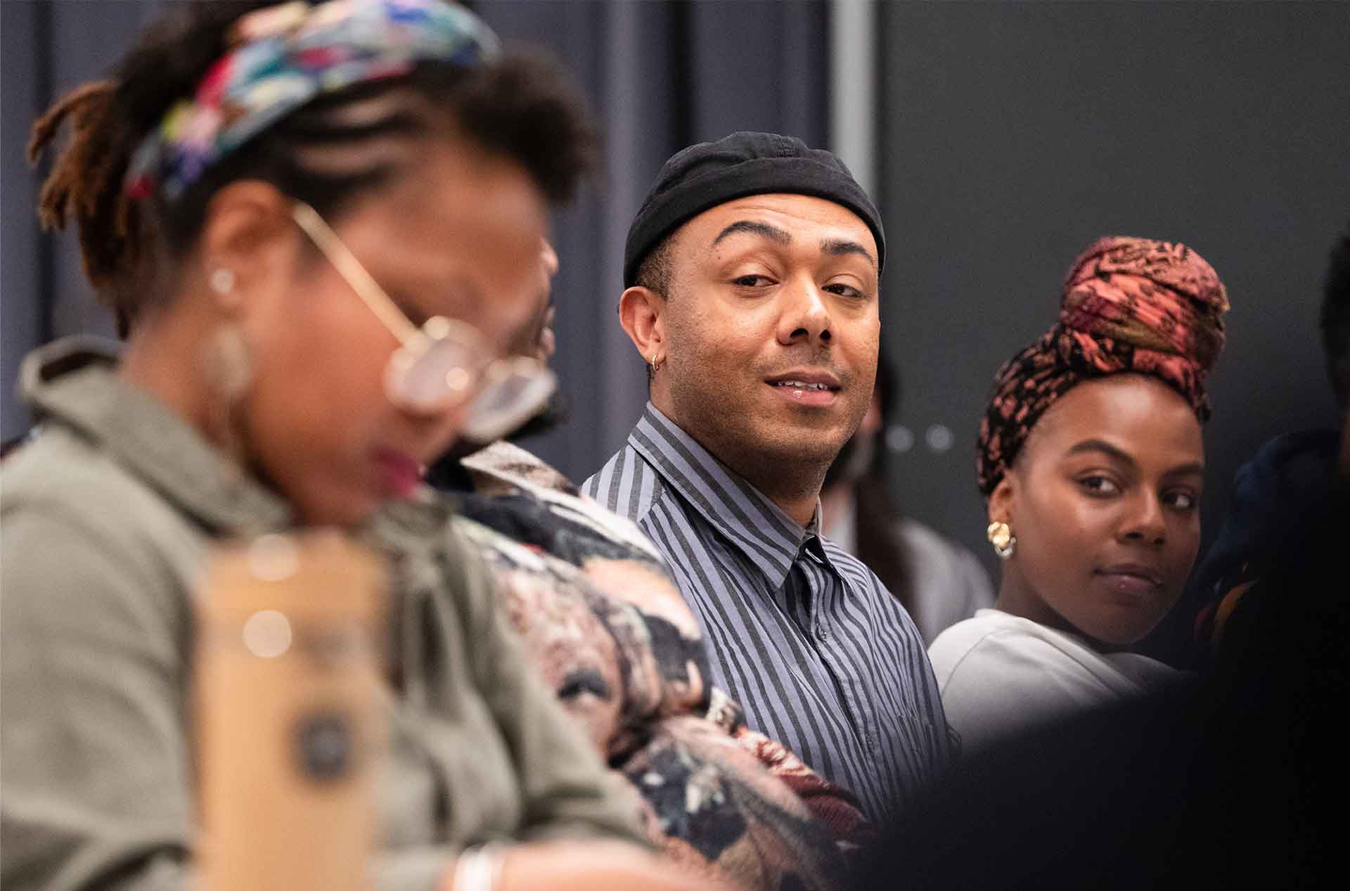 A person in a striped button-up and a black cap looks off to the side with a subtle smile. Two others in the foreground, slightly blurred, are also engaged in the moment.