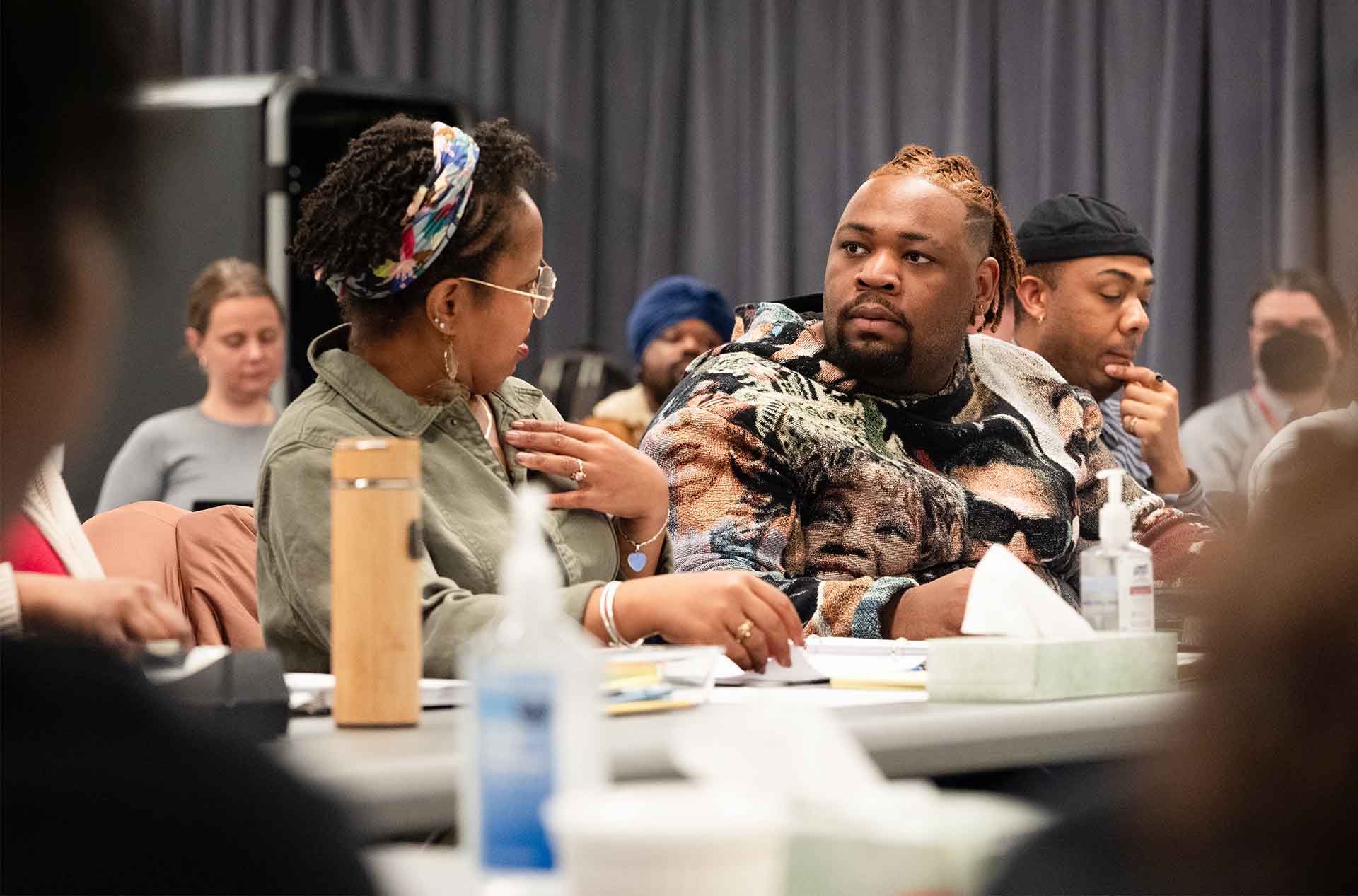 Two people sitting at a table engaged in conversation. One person wearing glasses and a headband gestures toward their chest, while the other, wearing a printed sweater, listens intently.