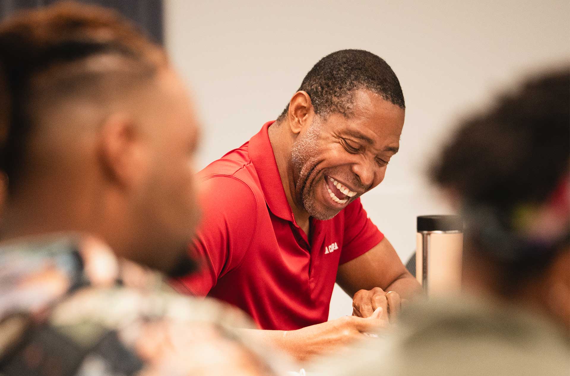 A person in a red polo shirt laughs while seated at a table, surrounded by others who are engaged in discussion. The foreground is blurred, focusing on their joyful expression.