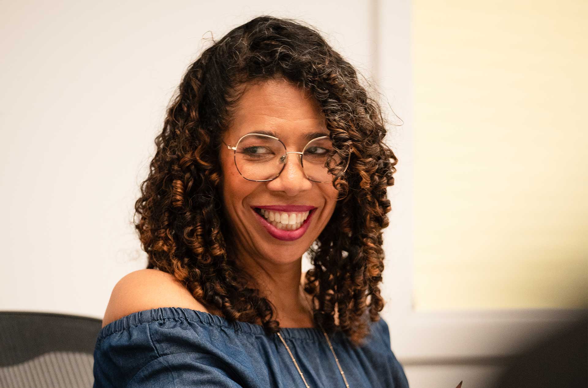 A person in a blue off-the-shoulder top and glasses smiles while sitting at a table. They appear to be engaged in a conversation, with open notebooks and papers in front of them.