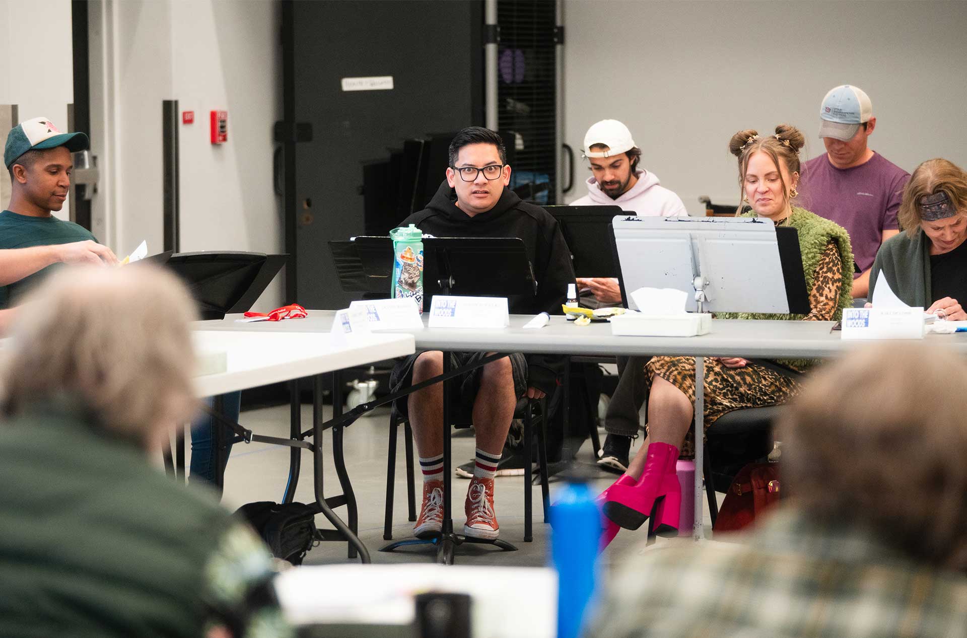 People seated at a table in a rehearsal room, reading scripts and engaged in discussion. One person wears bright pink platform boots, and nameplates and water bottles are visible on the table.