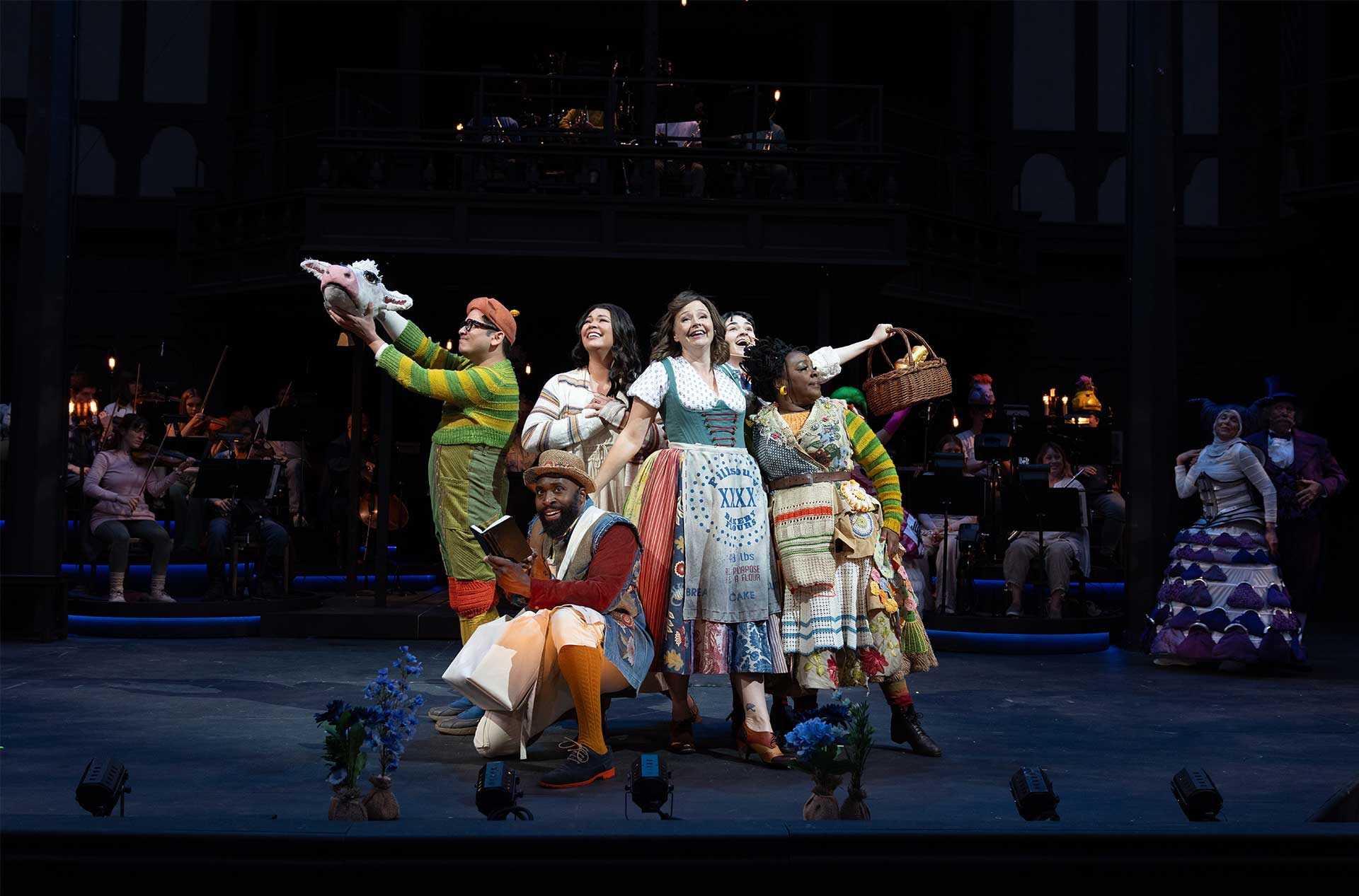 Six actors in colorful fairytale costumes pose joyfully at center stage, some holding props like a cow puppet head, book, and bread basket, with an orchestra and dark wood set in the background.