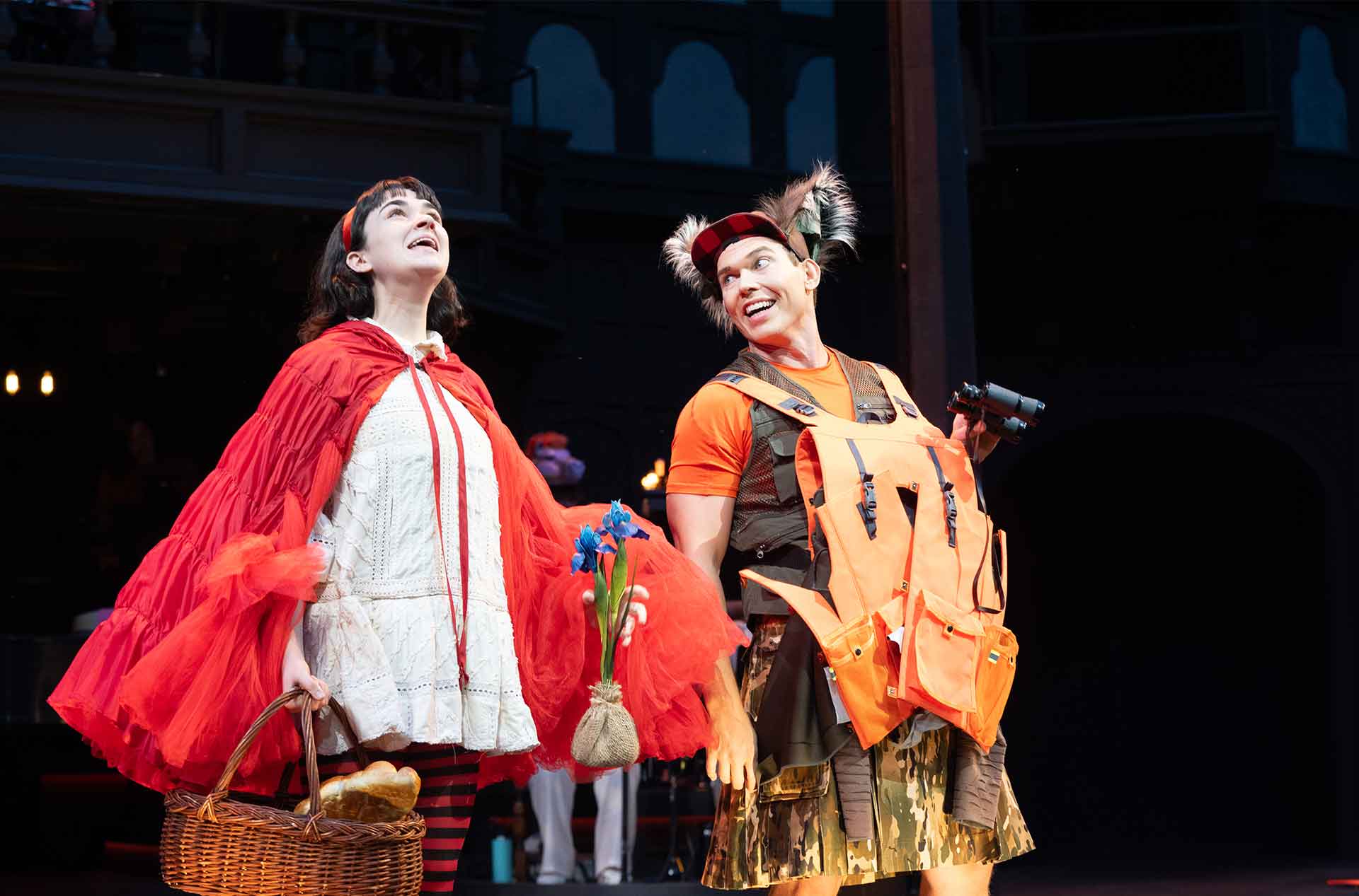 Two actors smile mid-performance; one dressed in a red cape and striped tights holding a basket and flowers, the other in camo and an orange vest with fuzzy ears and binoculars, suggesting a fairy tale encounter.