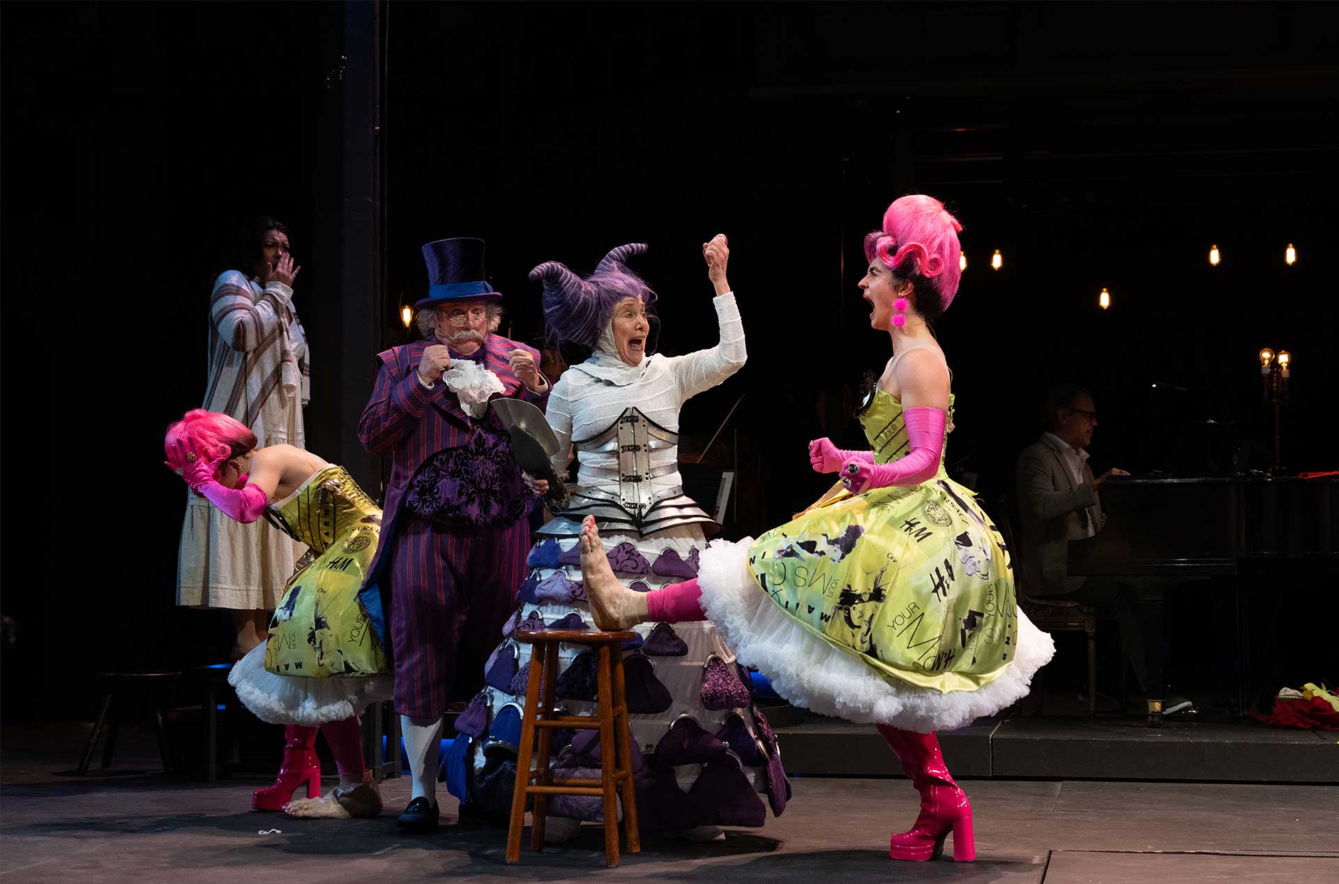 Five performers in eccentric costumes interact in an animated scene; one seated in a purple dress with curled horns, while another raises a leg in a lime green dress and pink gloves, mid-conversation.