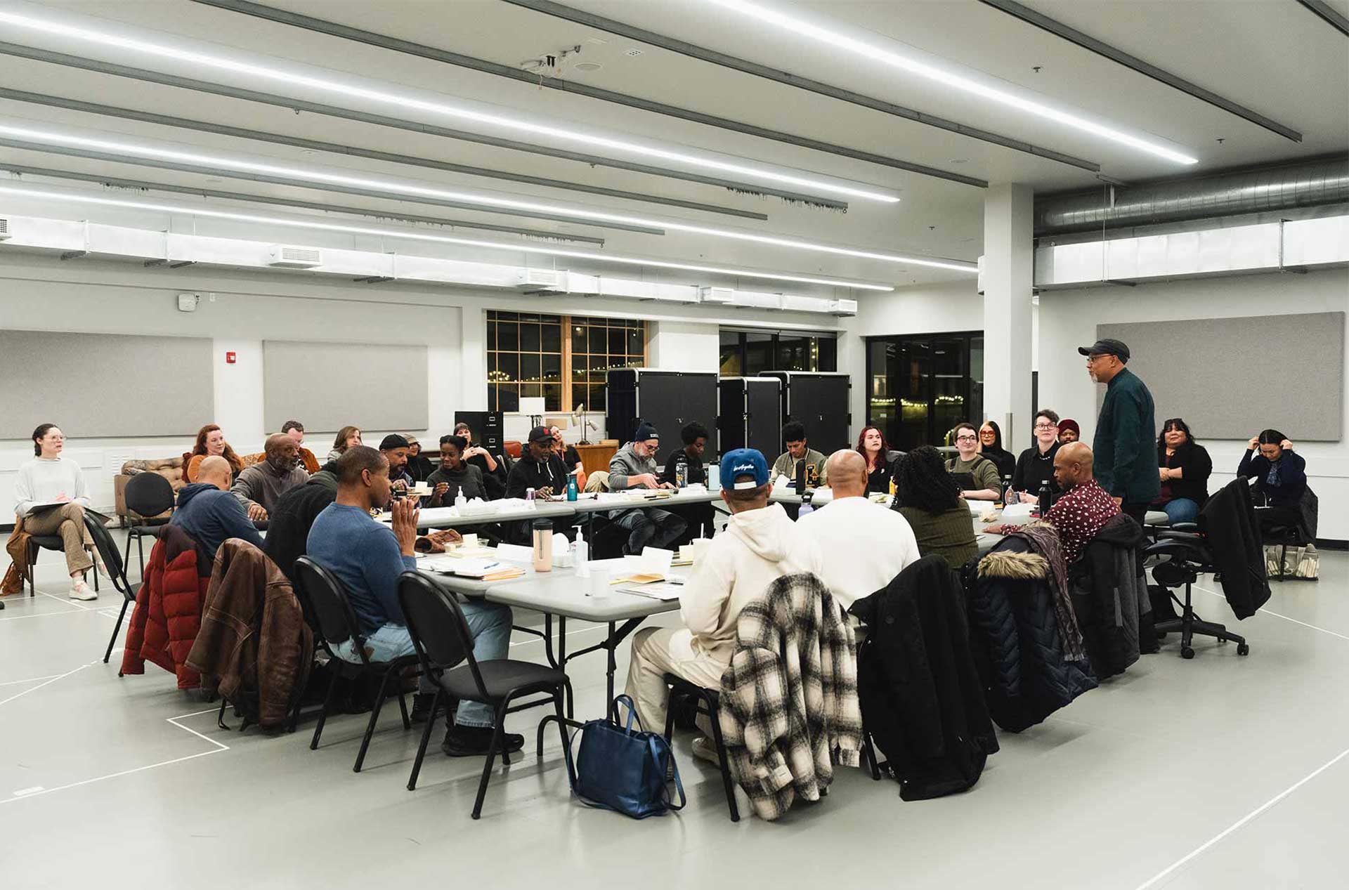 A large group of people seated around a U-shaped arrangement of tables in a brightly lit room, participating in a discussion. Some individuals take notes while others listen attentively.