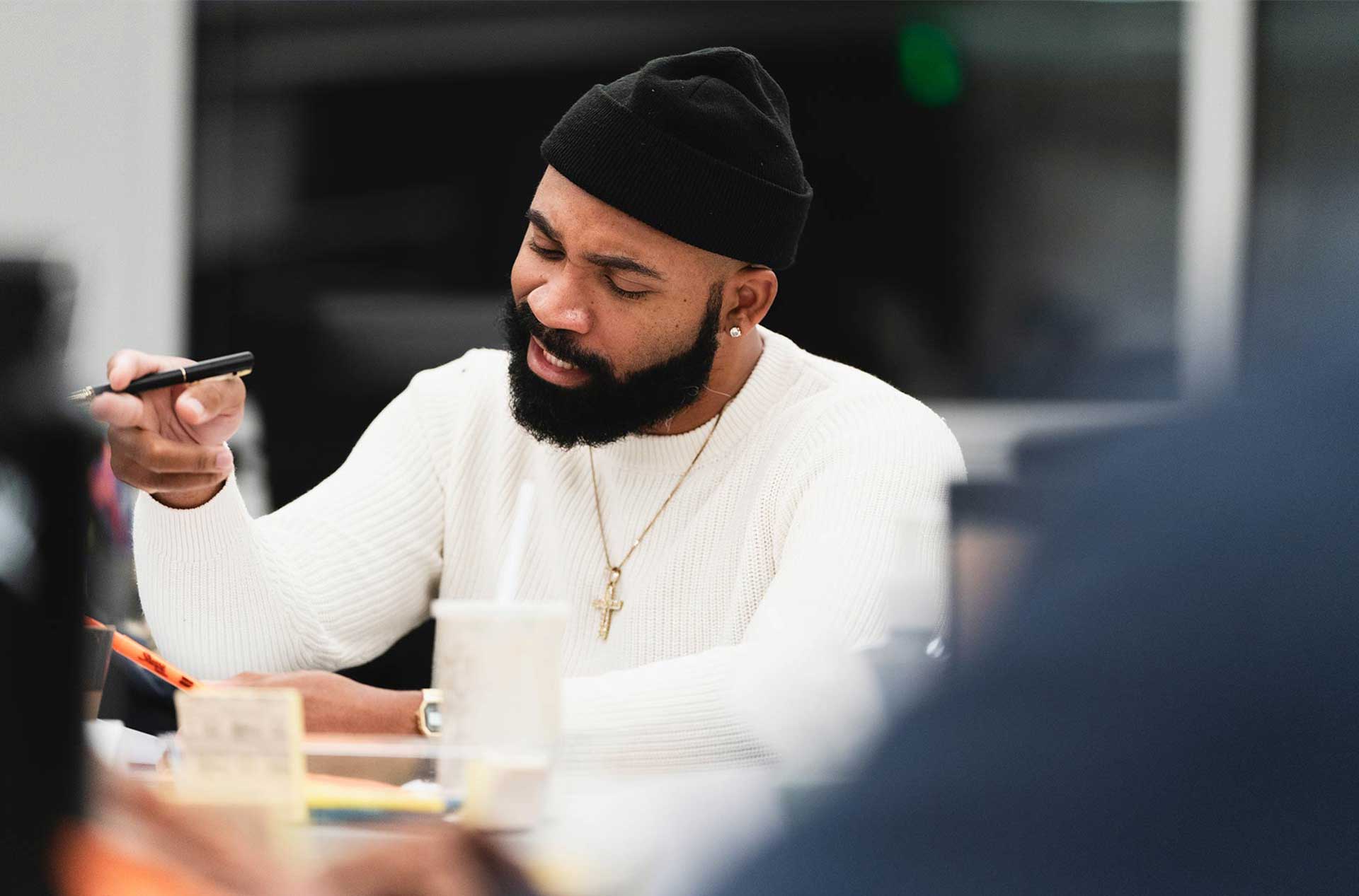 A person with a beard, wearing a white sweater and a black beanie, appears to be engaged in a discussion while holding a pen.