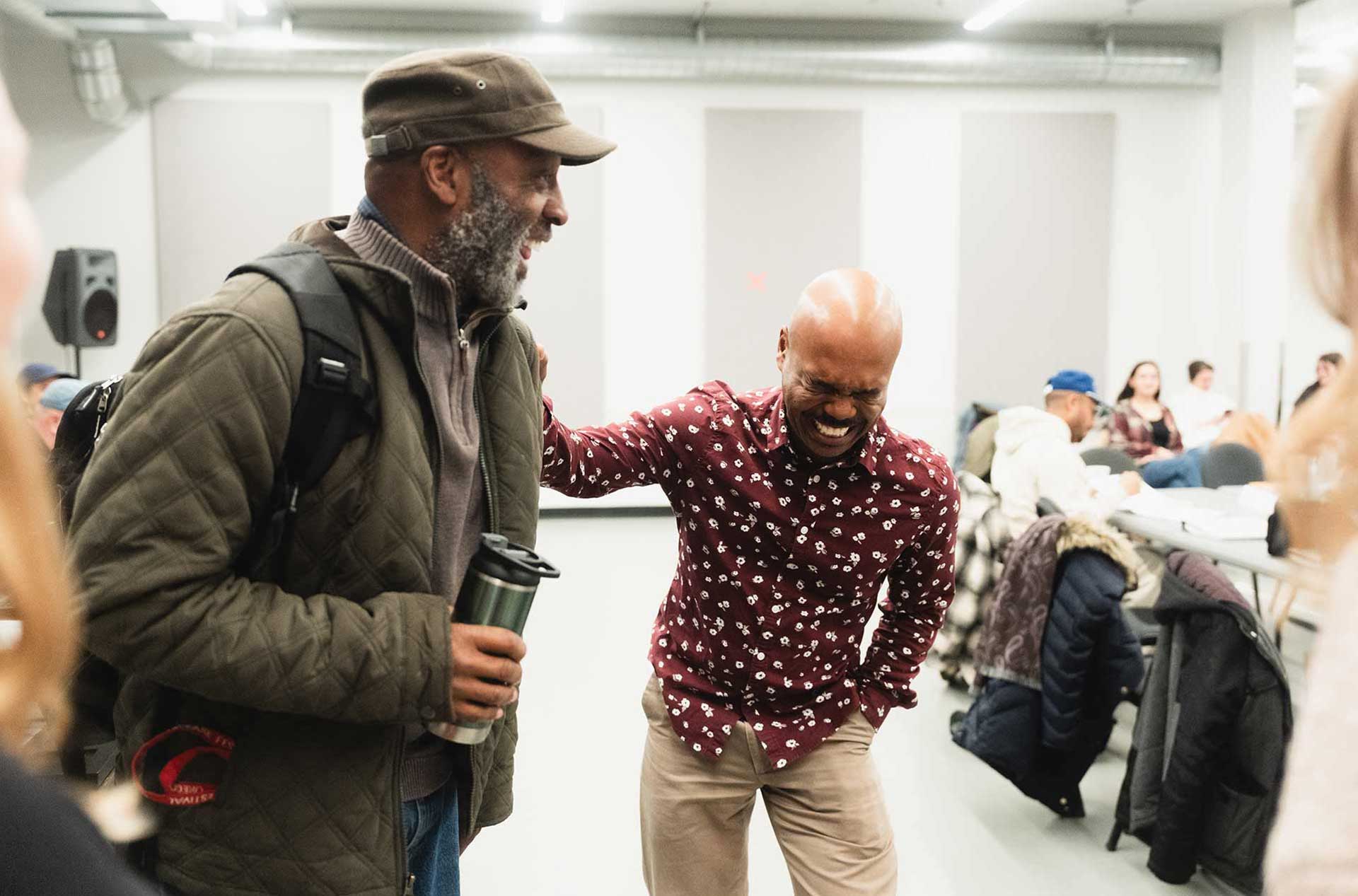 Two people standing together, laughing and chatting in a rehearsal.
