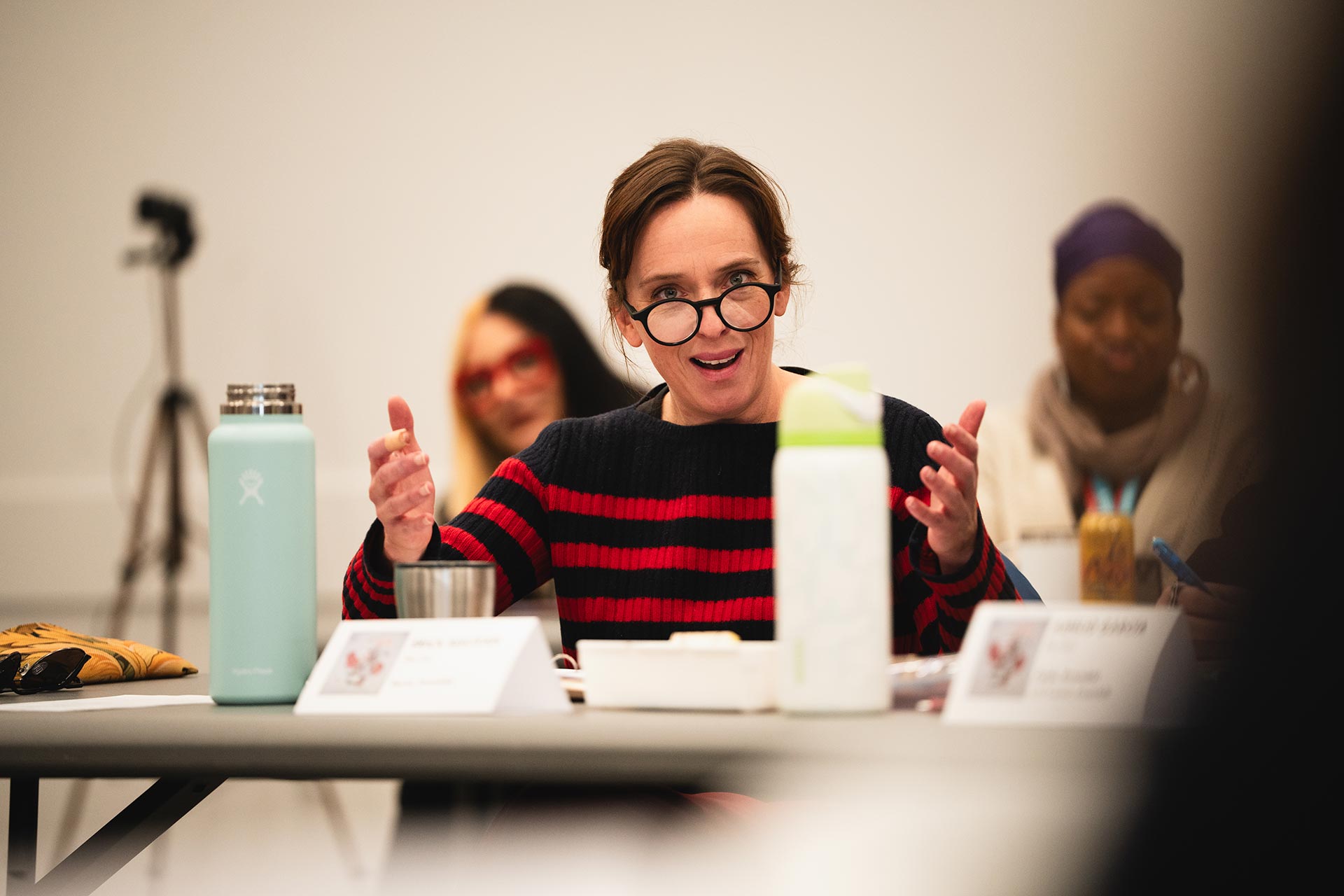 A person at the rehearsal table animatedly gesturing with both hands, engaging in a lively discussion. A nameplate and several water bottles are visible on the table in front of them.