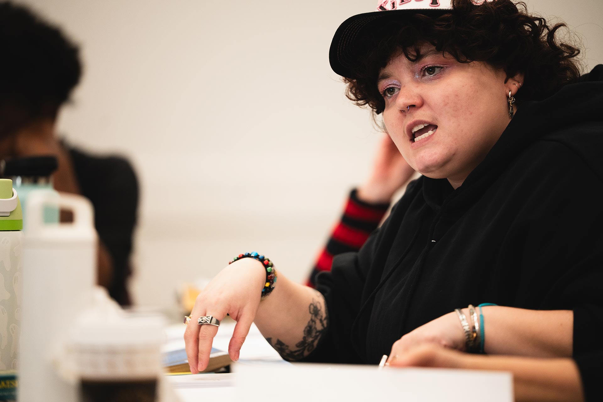 A rehearsal participant speaks passionately, gesturing with one hand while seated at a table covered with scripts and water bottles. They are wearing a hoodie and a cap with lettering, expressing an idea during the discussion.