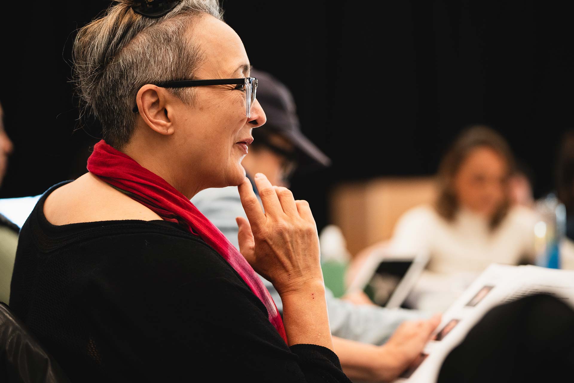 A creative team member or cast participant smiles thoughtfully during the rehearsal. They are holding a script and wearing glasses, with a red scarf draped around their neck.
