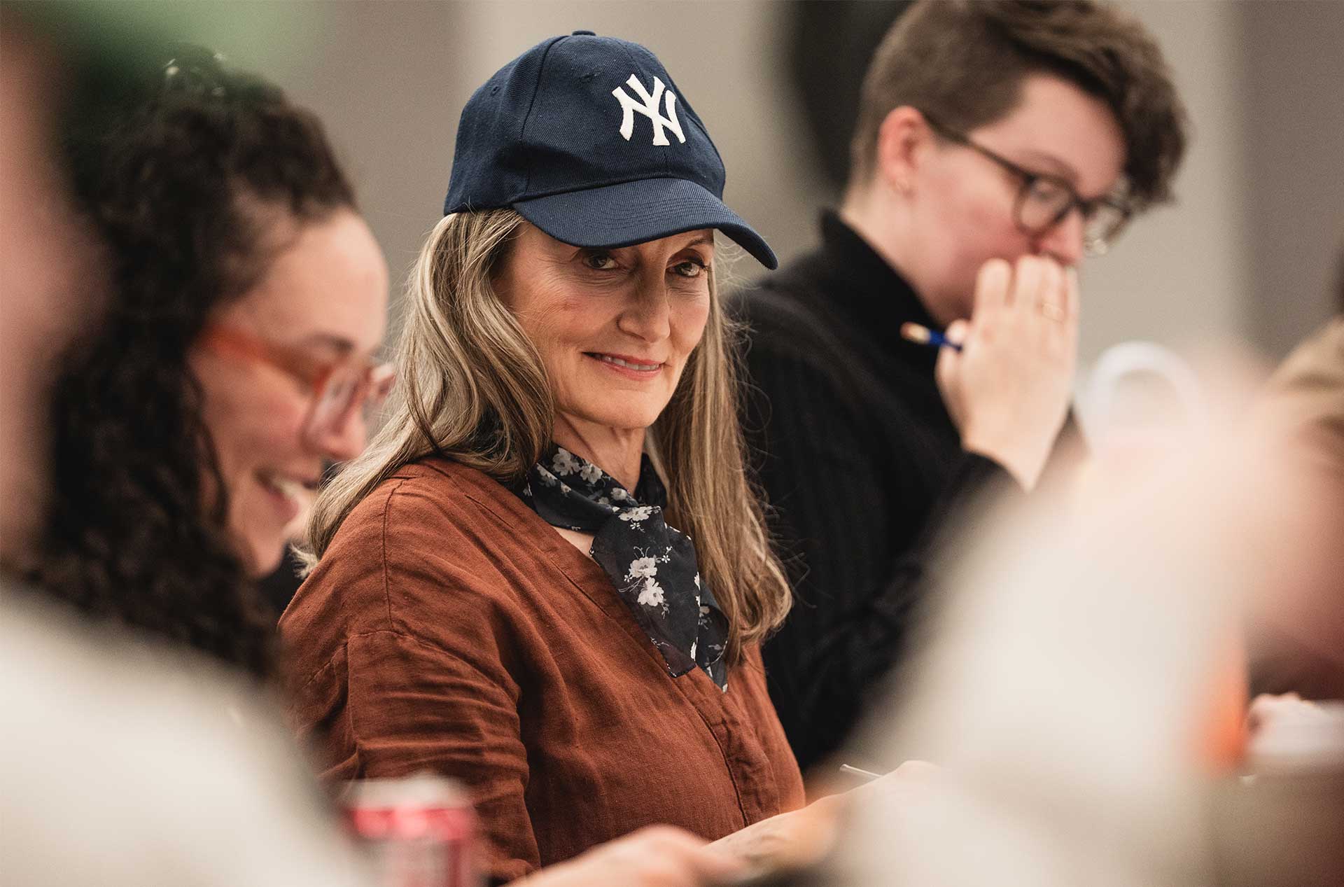 Person wearing a New York Yankees baseball cap and a floral scarf looks ahead with a slight smile, seated at a table during a script read-through.