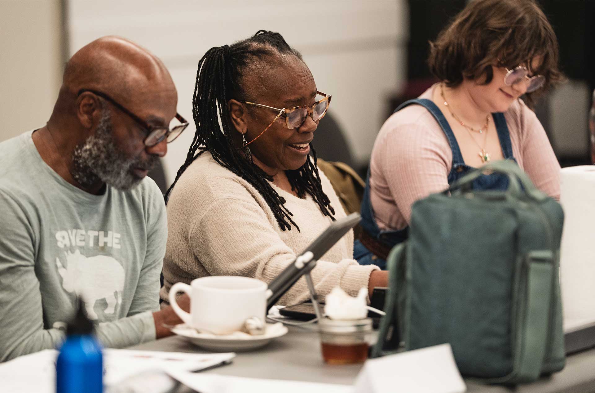 Three people seated at a long table read and discuss scripts; mugs, bags, and bottles are scattered across the table.