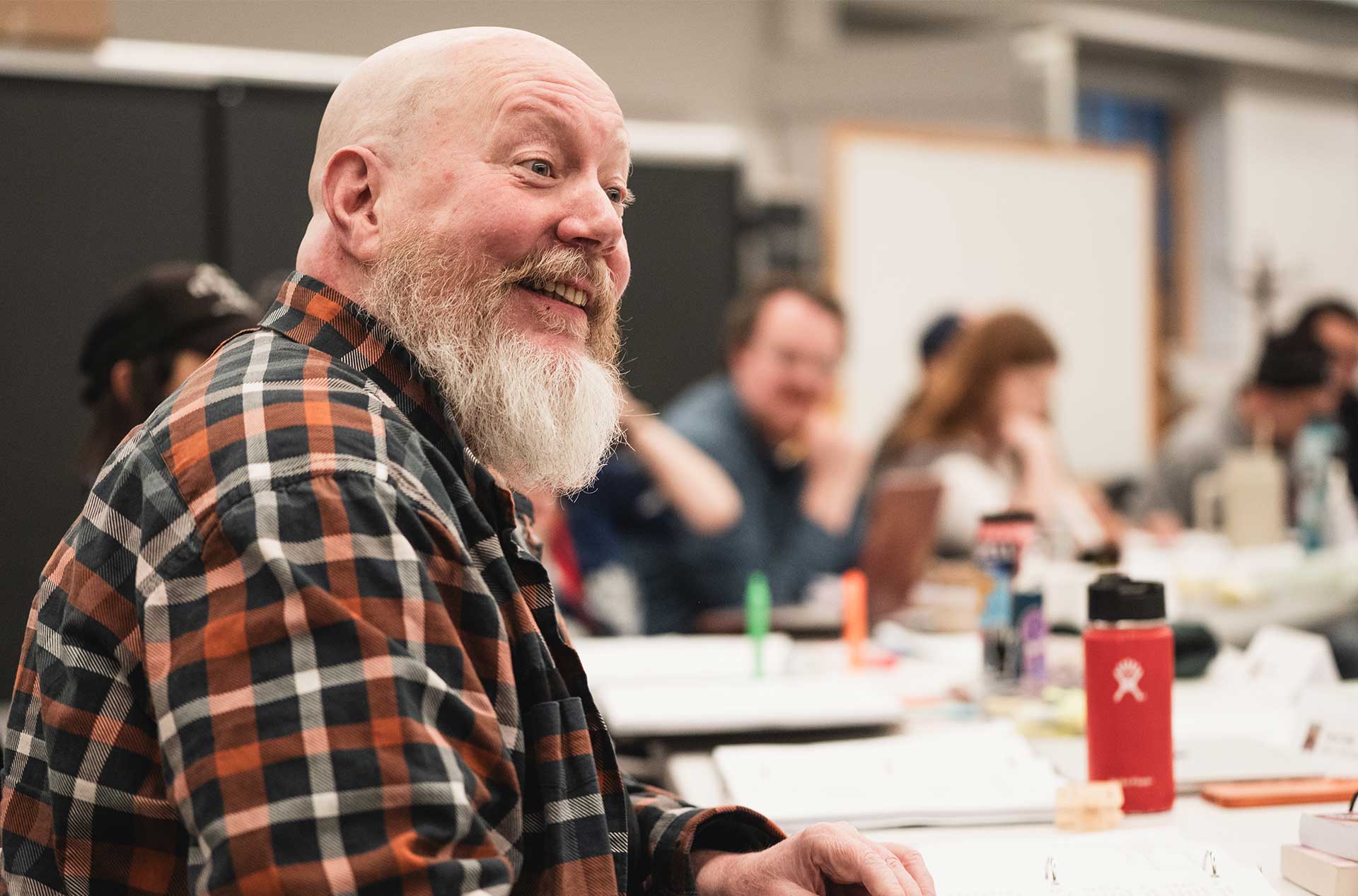 A person with a full white beard and plaid shirt smiles warmly while seated at a long rehearsal table filled with papers and water bottles.