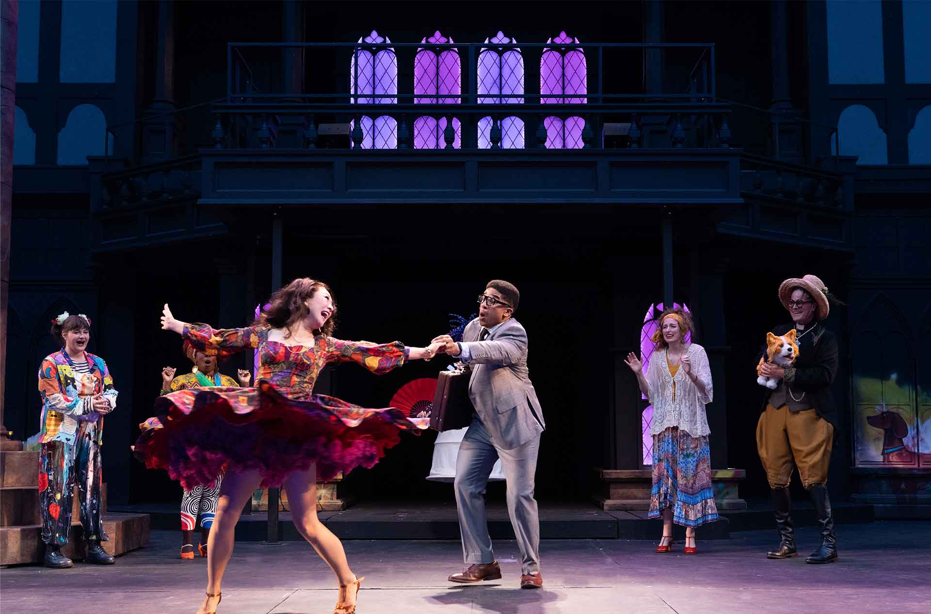 An actor in a swirling, colorful dress twirls energetically while holding hands with another in a grey suit, who appears surprised. Others in whimsical outfits, including one holding a plush corgi, cheer them on.