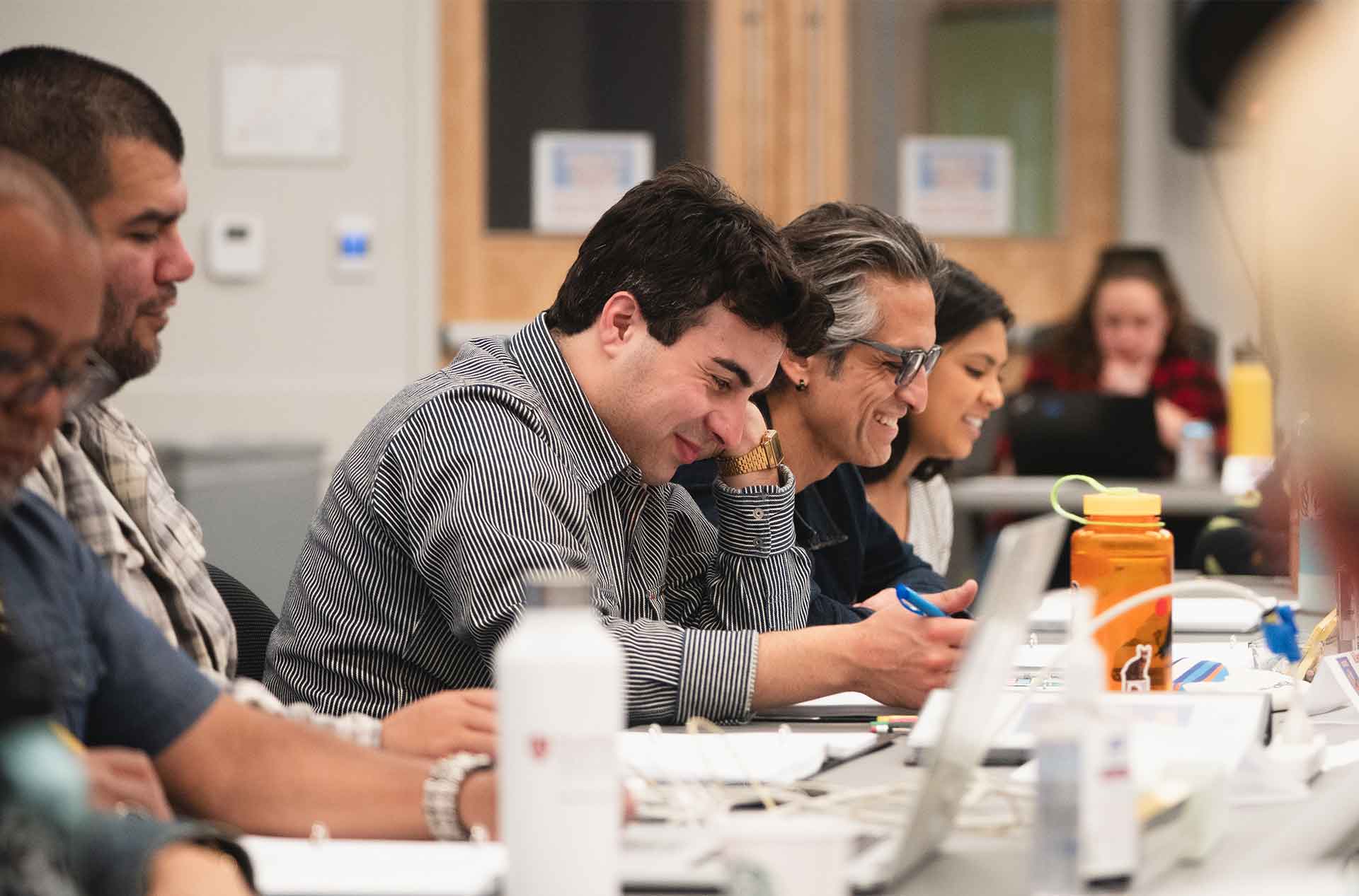 Participants seated at a table during a rehearsal, smiling and writing notes, with papers, laptops, and water bottles in front of them.